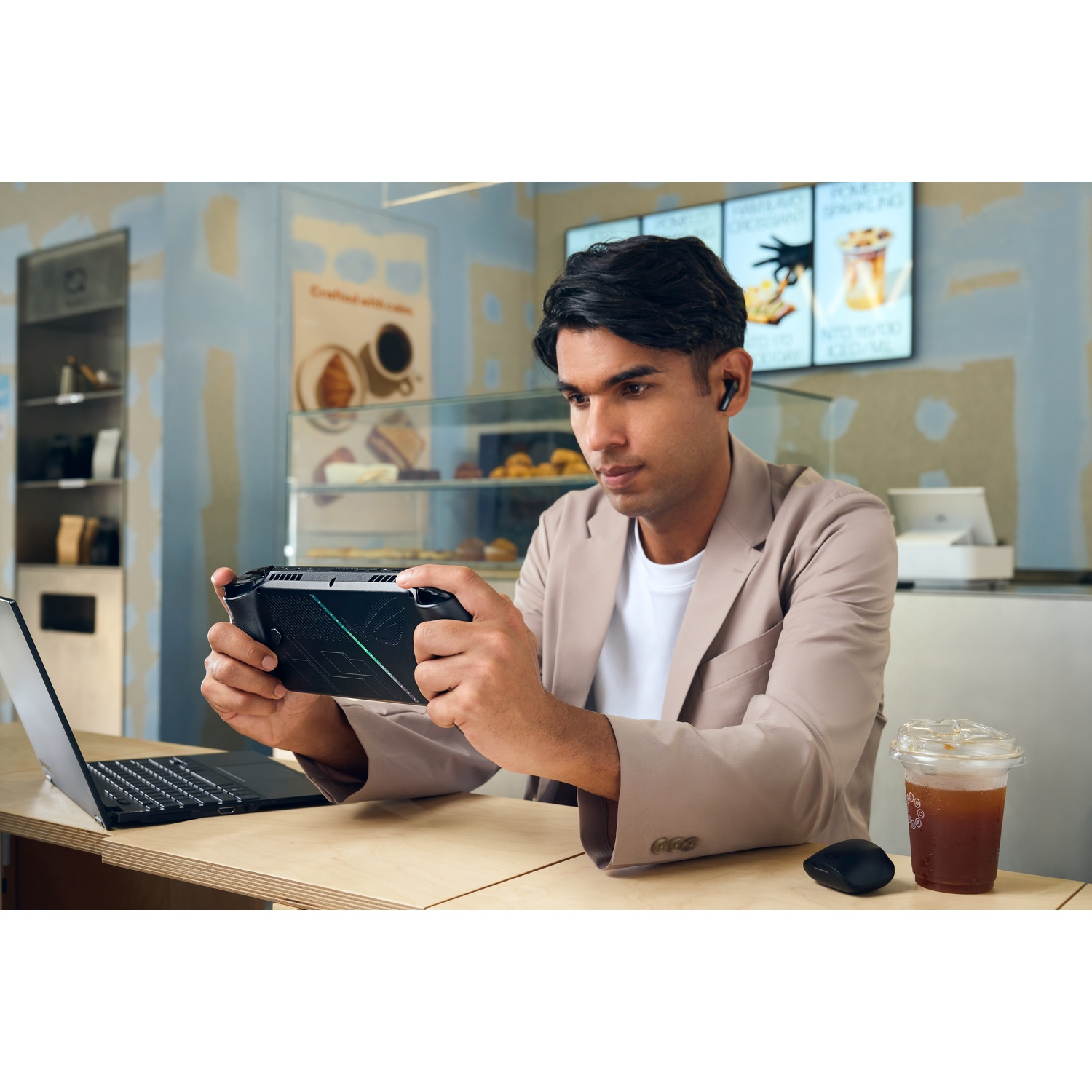 Un uomo con auricolari gioca con un dispositivo di gioco portatile su un tavolo da caffè. Sul tavolo ci sono anche un laptop e un caffè freddo.