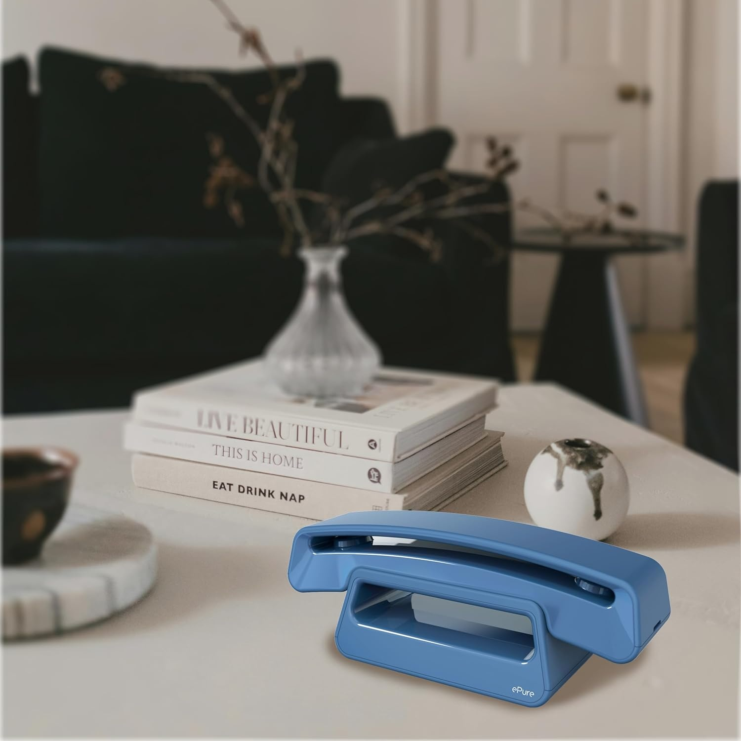 Un telefono ePure blu è appoggiato su un tavolo bianco con libri e decorazioni. Un divano scuro è sullo sfondo.
