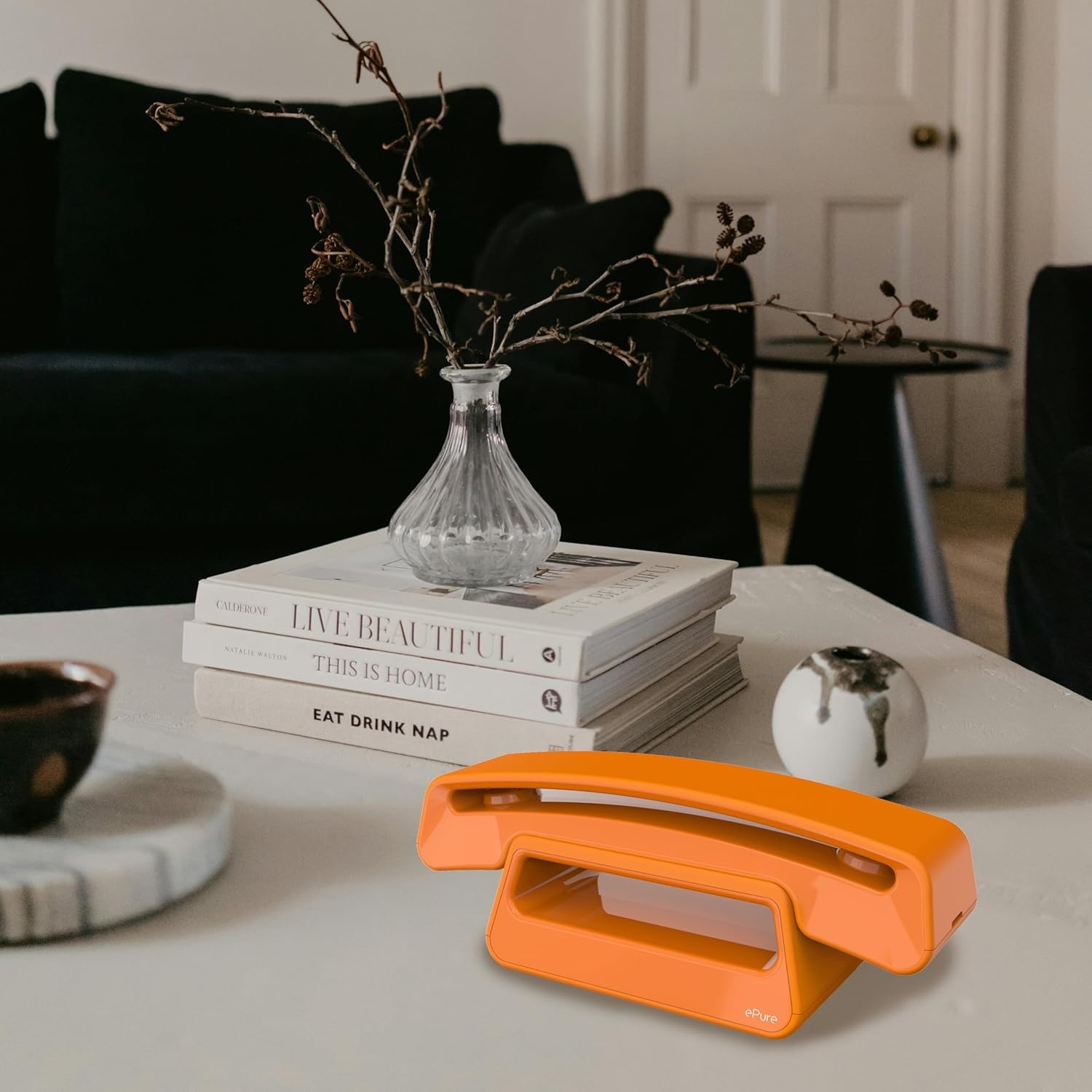 Un telefono arancione è su un tavolo bianco con libri e un vaso. Un divano nero e una porta sono sullo sfondo sfocato.