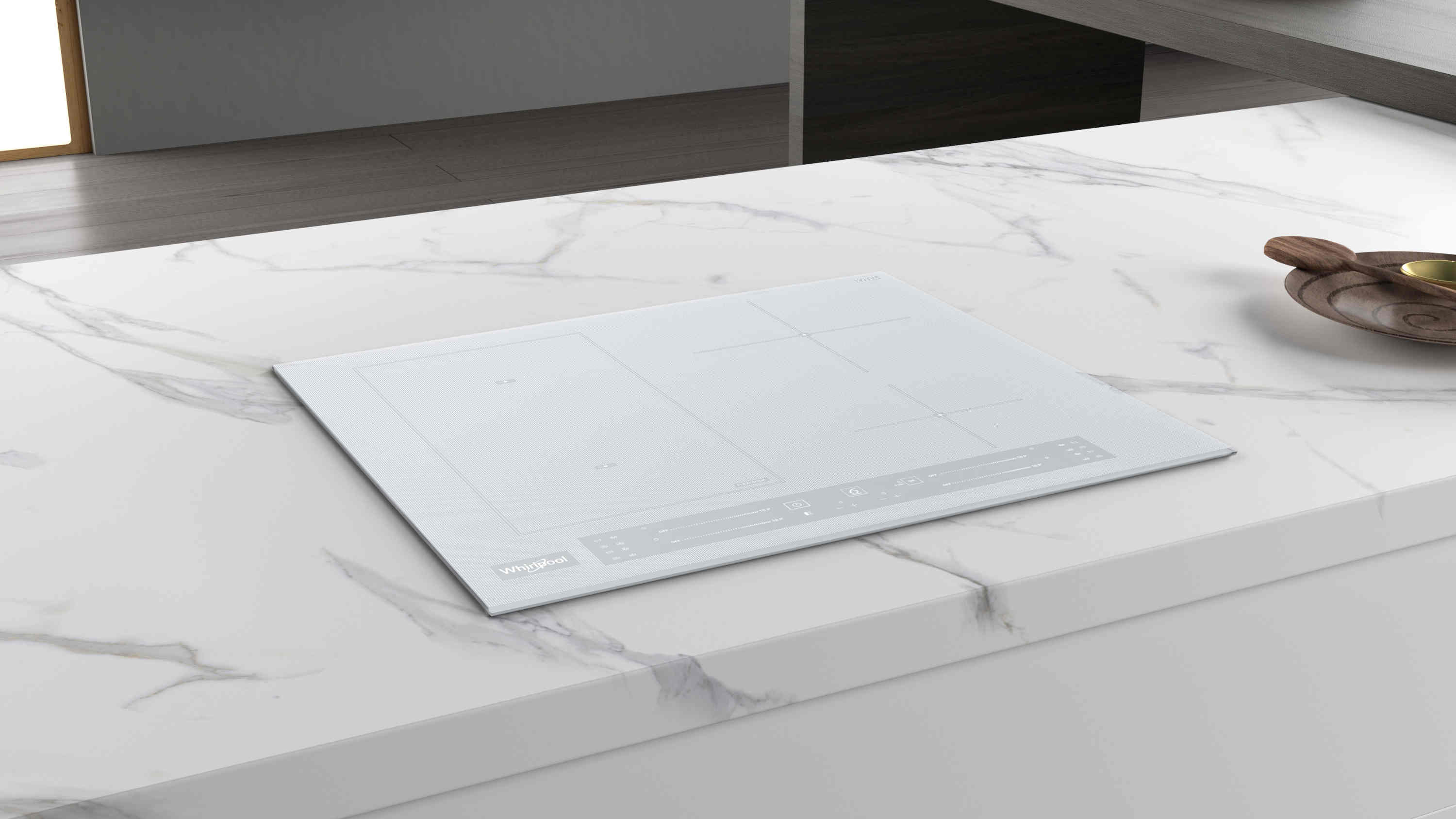 Biała płyta indukcyjna Whirlpool na białym marmurowym blacie. Drewniany talerz z łyżką w tle.