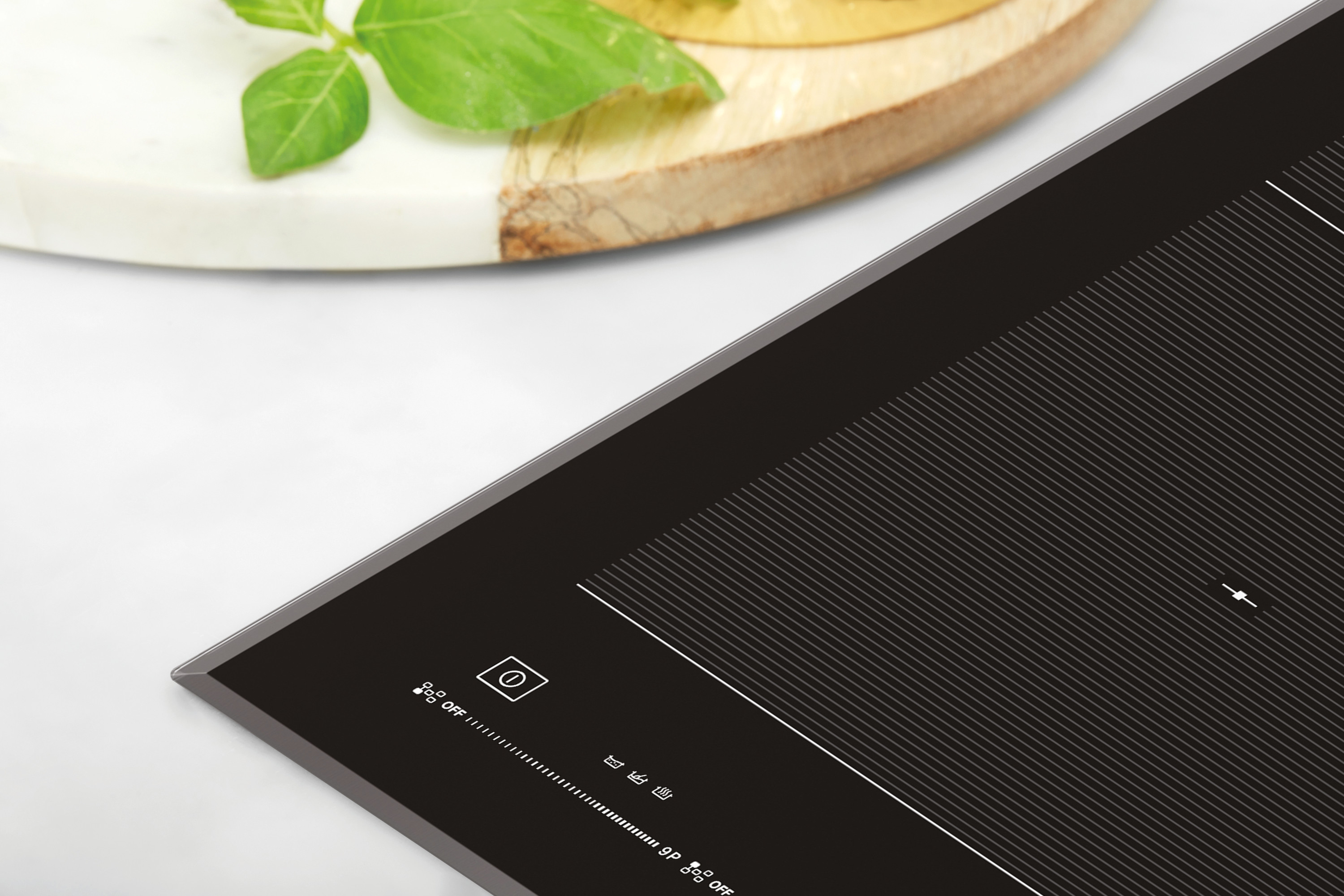 Zbliżenie czarnej płyty indukcyjnej z białymi liniami i panelem sterowania. Drewniana deska z liśćmi bazylii na górze.
