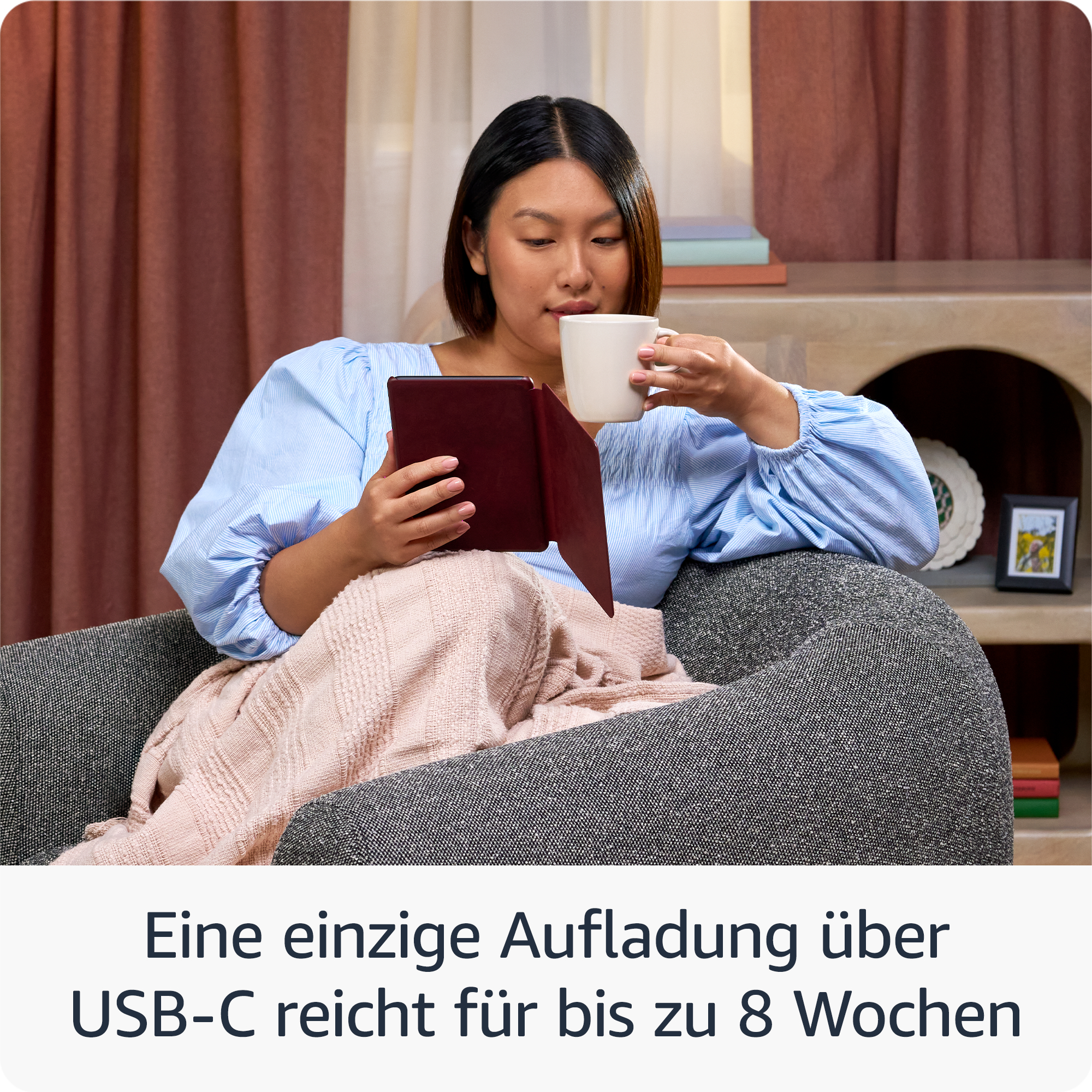 Frau liest Tablet, trinkt aus einem Becher und sitzt auf einem Stuhl. Hintergrund: Decke, Vorhang und Dekoration.