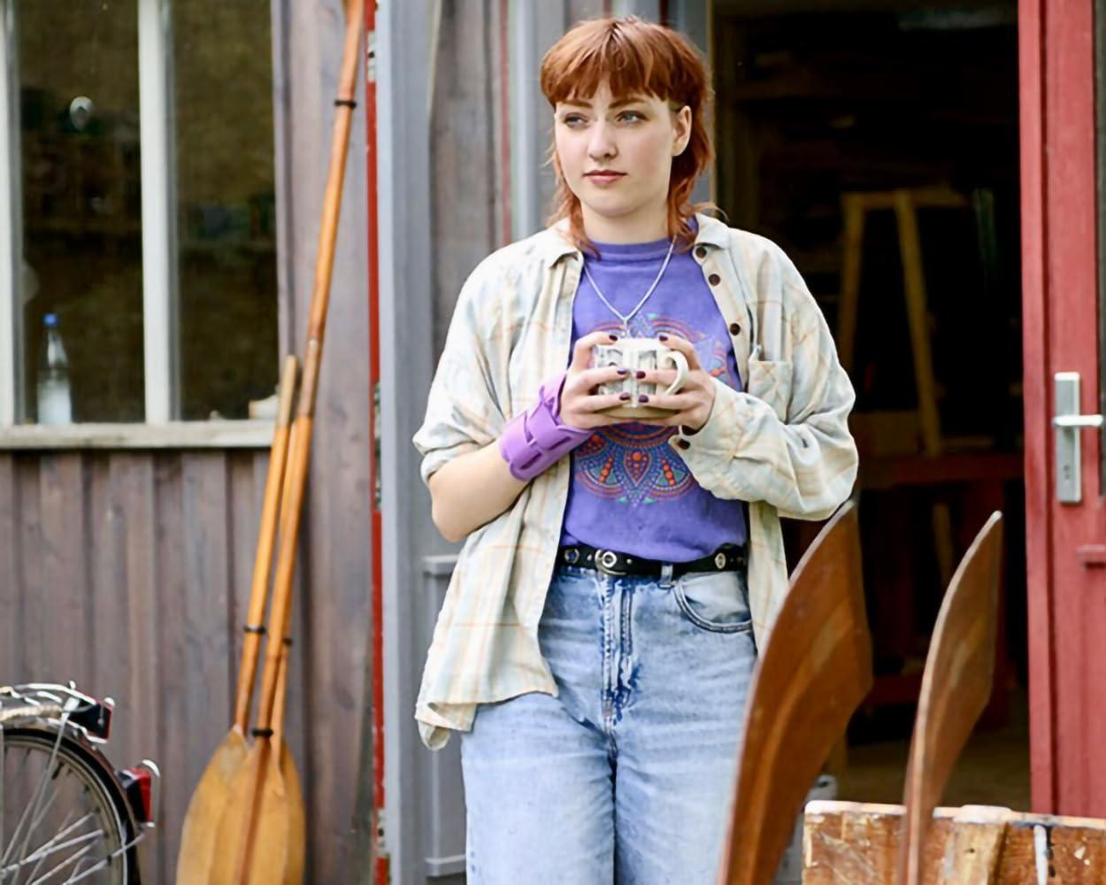 Eine Frau mit roten Haaren hält einen Becher. Sie trägt ein lila Hemd und Jeans und steht an einer Holzkonstruktion mit Rudern.