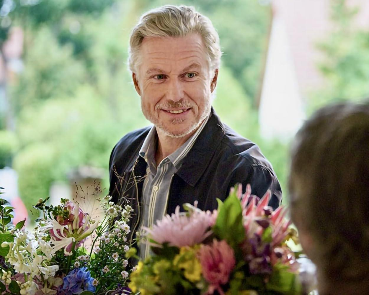 Ein lächelnder Mann mit grauem Haar hält einen Strauß bunter Blumen. Er trägt eine dunkle Jacke und ein gestreiftes Hemd. Eine Person ist rechts.