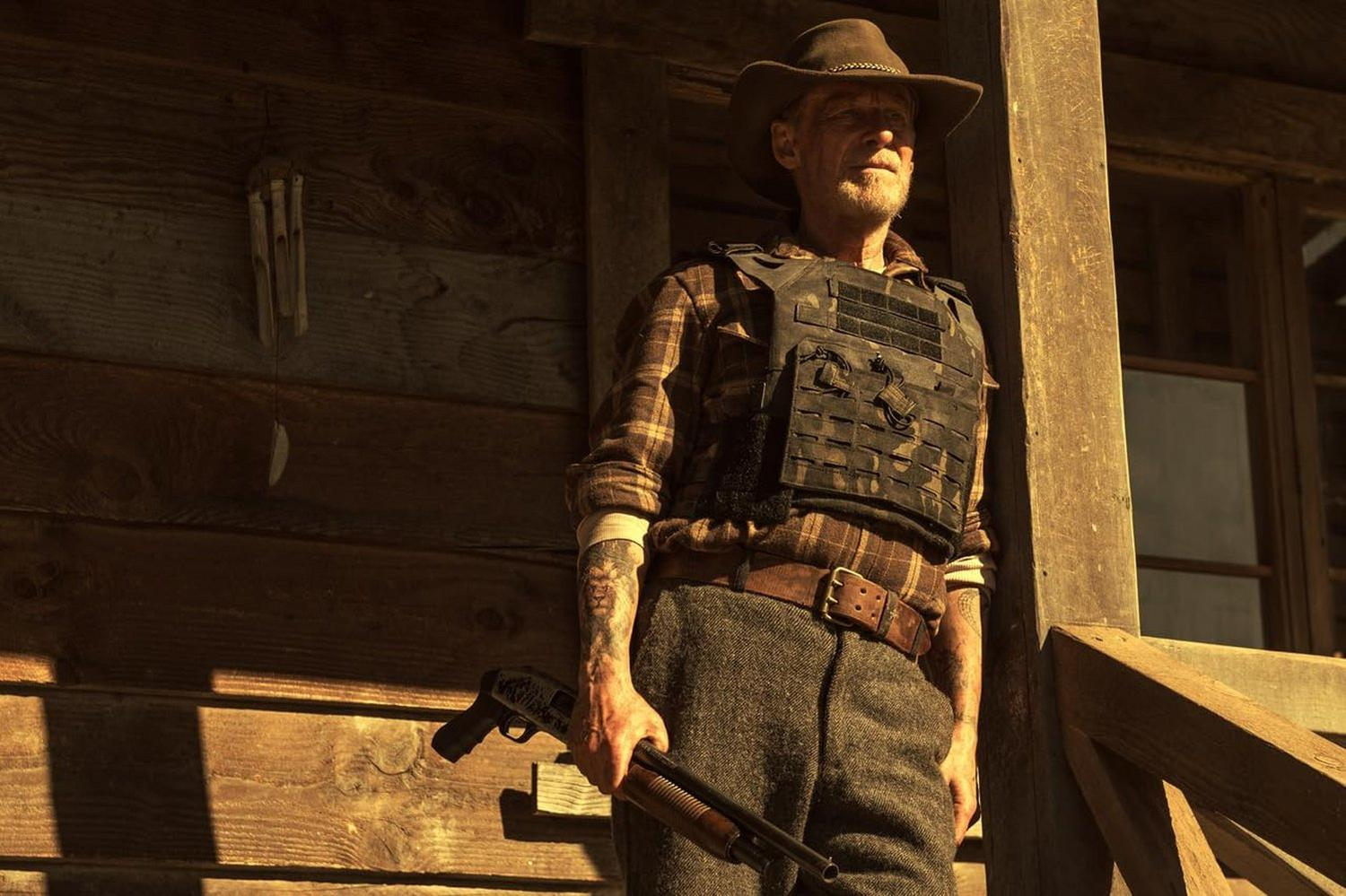 Mann mit Cowboyhut und taktischer Weste hält eine Schrotflinte. Er steht an einem Holzgebäude, mit Holz-Windspielen an der Wand.