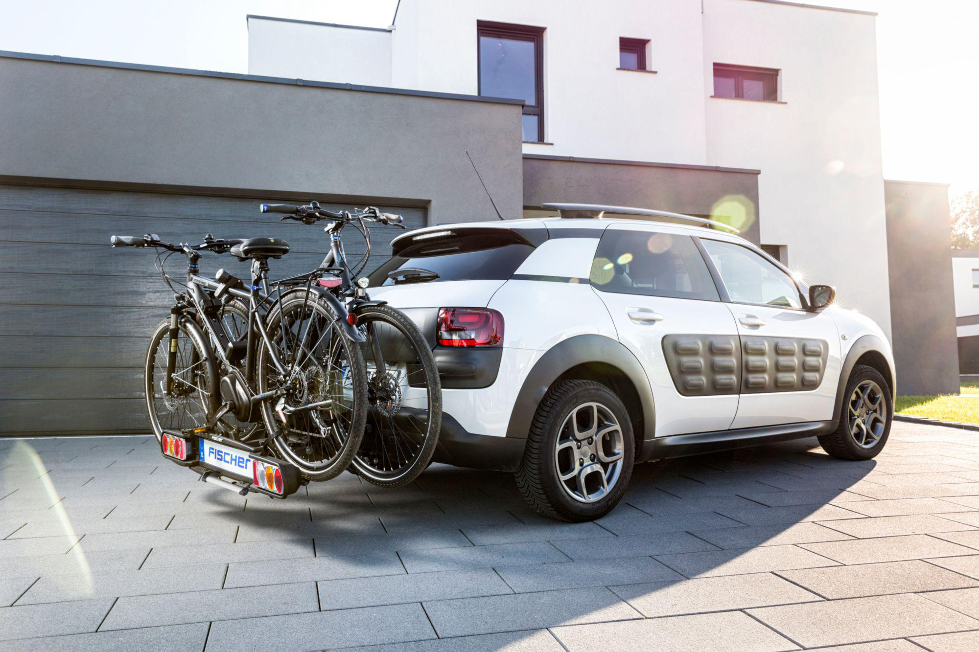 Weißes Auto mit zwei Fahrrädern hinten. Das Auto steht auf einer Backsteinauffahrt vor einem Haus.