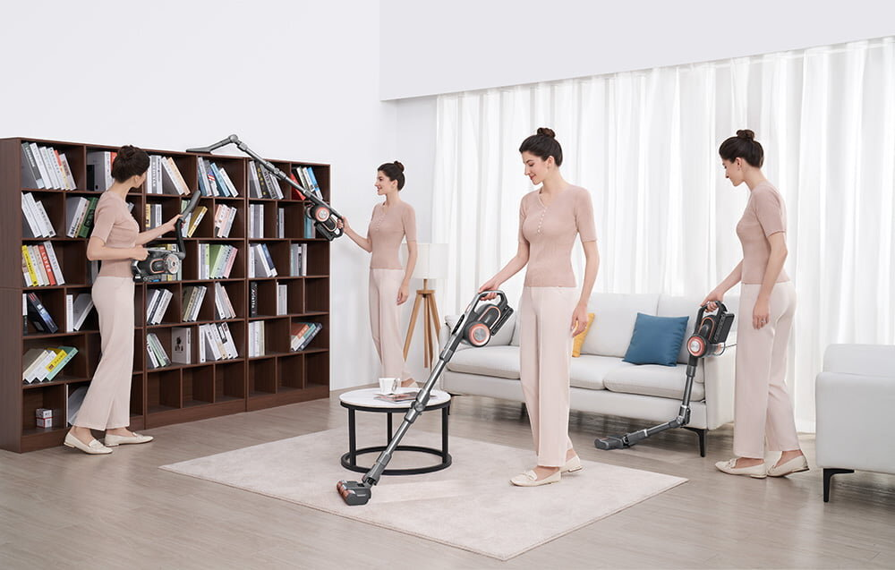 Kobieta odkurza regał, dywan i podłogę w salonie. Druga kobieta odkurza. Biała sofa, stolik kawowy i zasłony w tle.