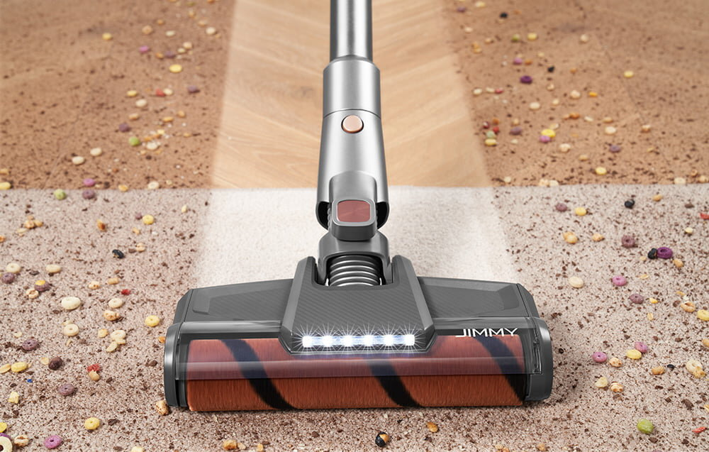 Srebrny odkurzacz czyści dywan z kawałkami płatków śniadaniowych. Oczyszczony obszar jest jaśniejszy. Odkurzacz ma markę JIMMY.