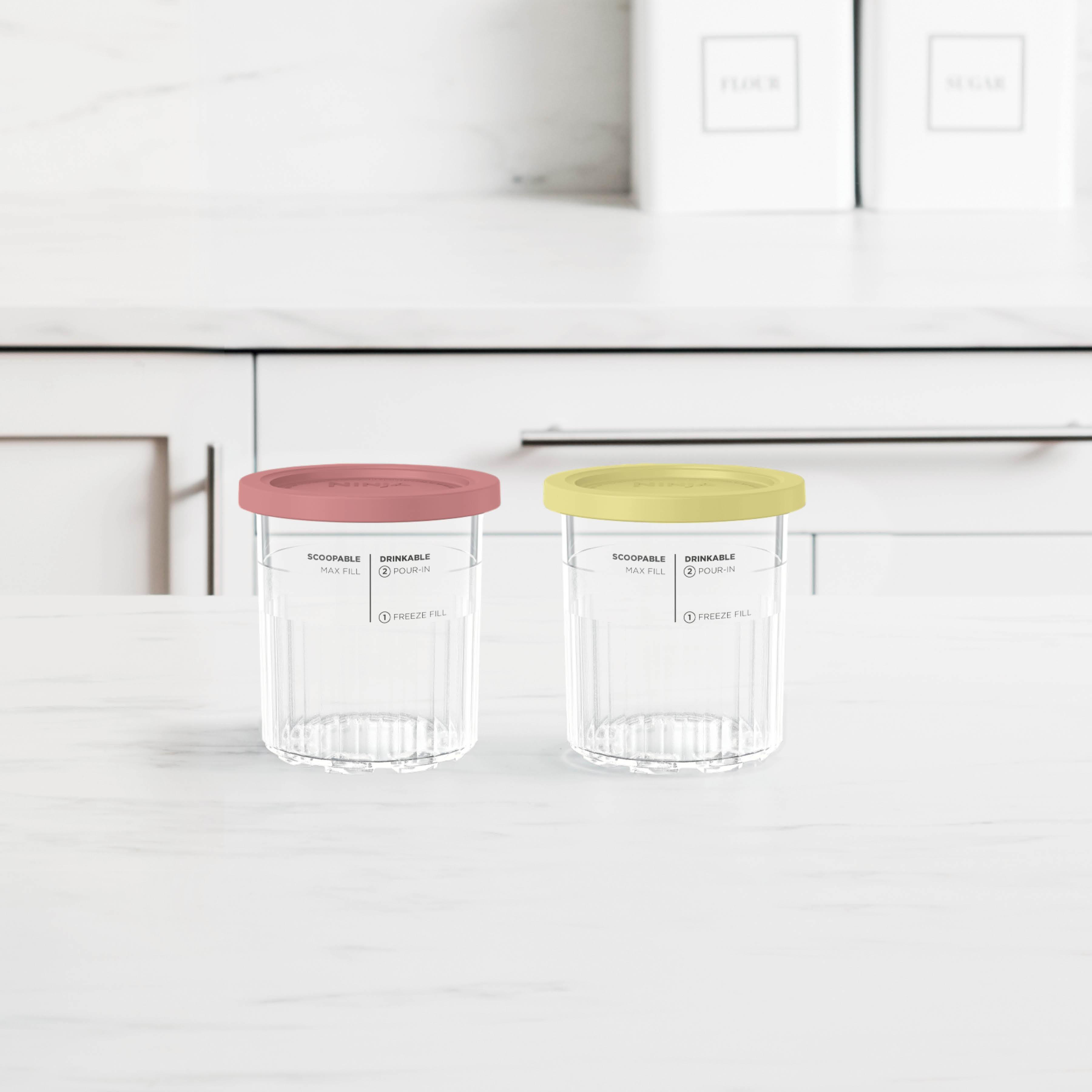 Zwei klare Plastikbecher mit rosa und gelben Deckeln stehen auf einer weißen Oberfläche. Etiketten zeigen Füllstände an.