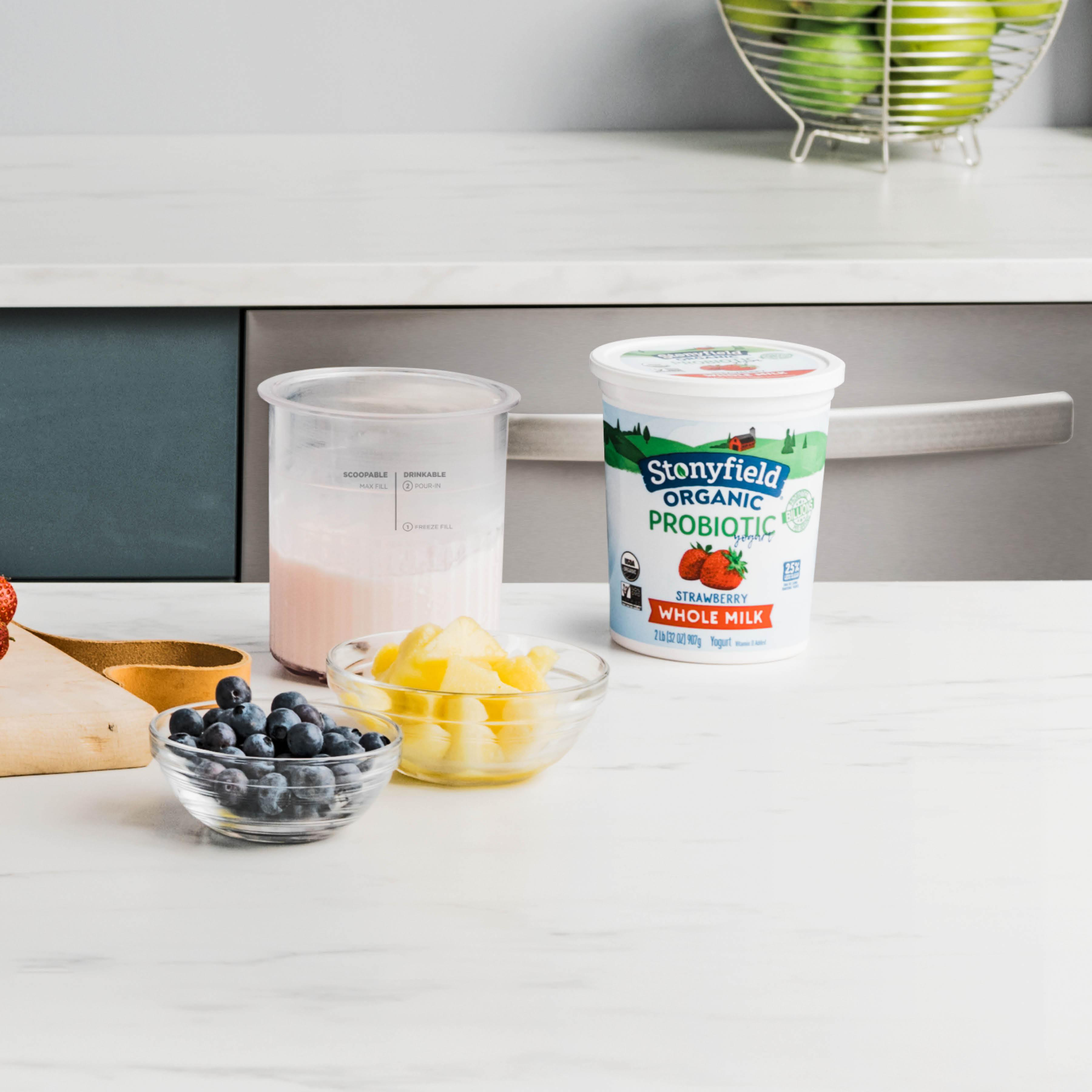 Ein Tisch mit Obst, Joghurt und einem Messbecher. Eine Schüssel mit Blaubeeren und Ananas ist vorhanden.