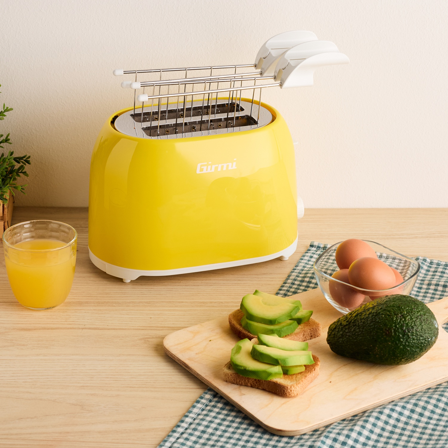 Tostapane giallo con toast di avocado, uova e succo d'arancia su un tavolo di legno. Sono presenti anche un avocado intero e un tovagliolo a scacchi.