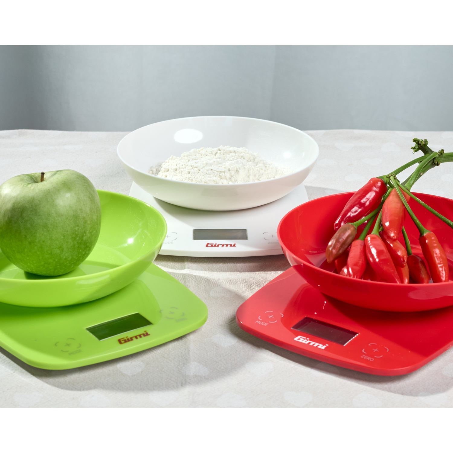 Tre bilance da cucina con ingredienti. Una mela verde poggia su una bilancia verde, farina su una bilancia bianca e peperoni rossi su una bilancia rossa.