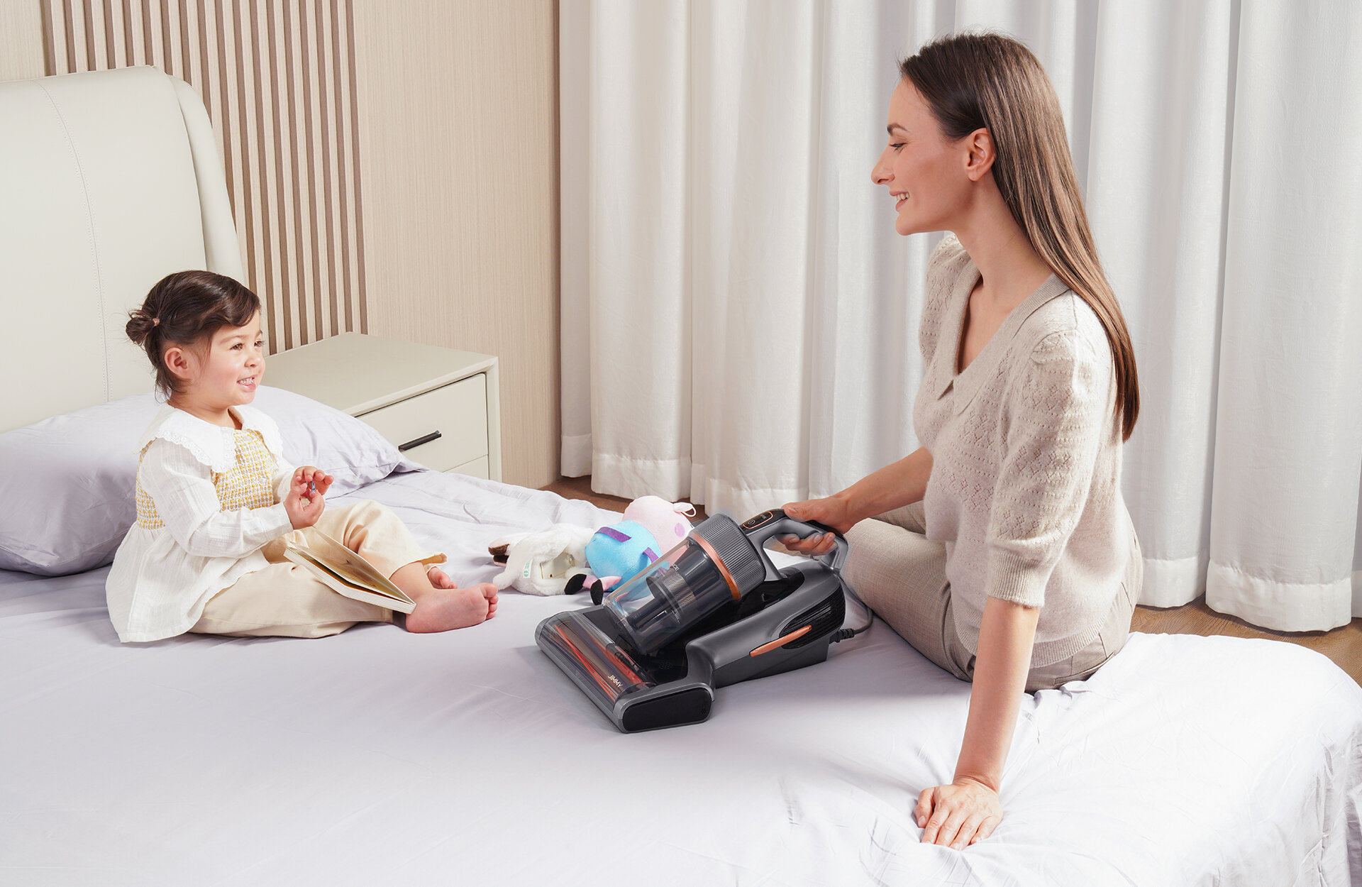 Kobieta i dziecko na łóżku. Odkurzacz jest umieszczony na łóżku między nimi. Kobieta uśmiecha się do dziecka.