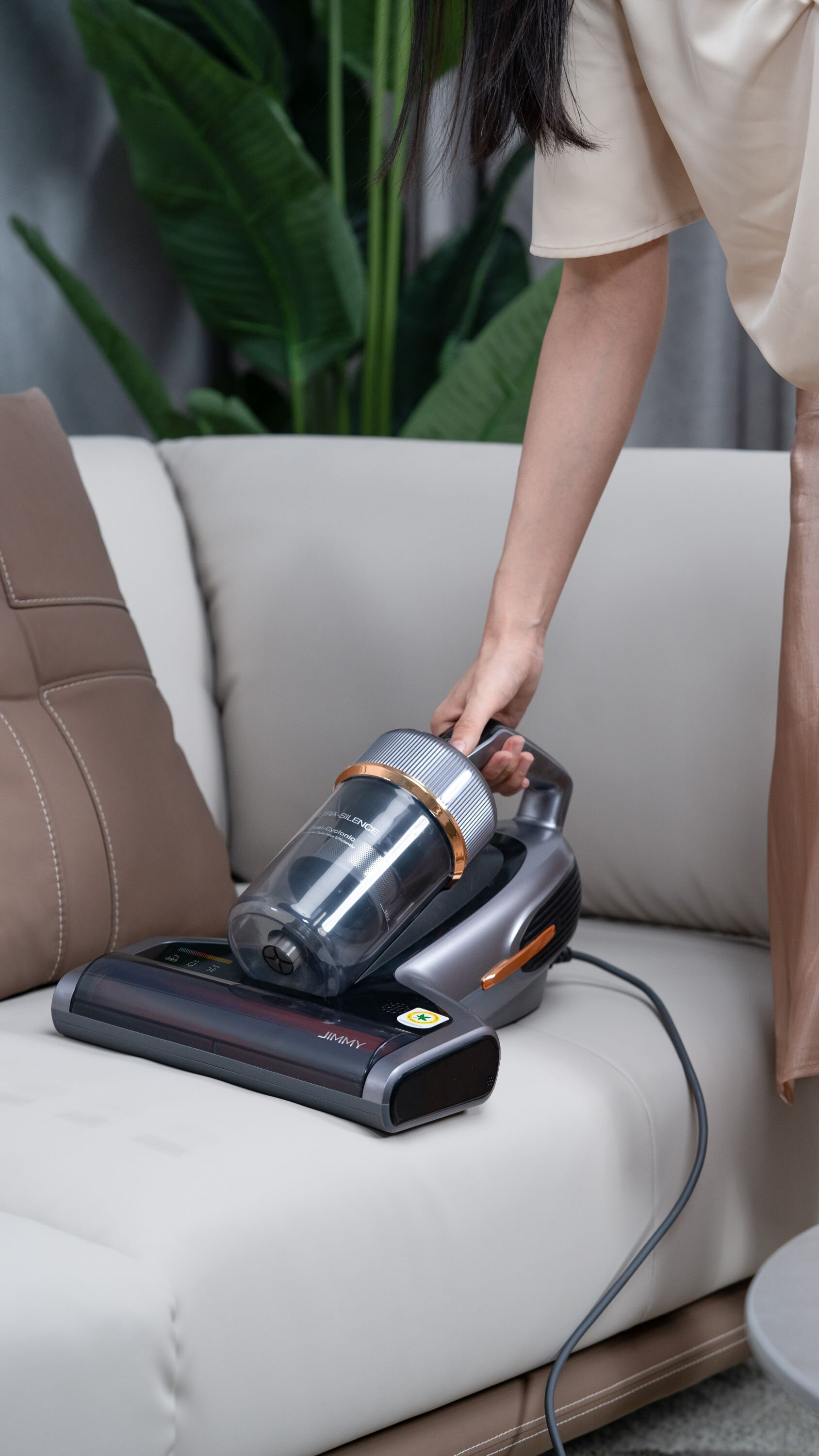 Osoba trzyma odkurzacz na kanapie. Odkurzacz jest czarny i szary. Zielona roślina w tle.