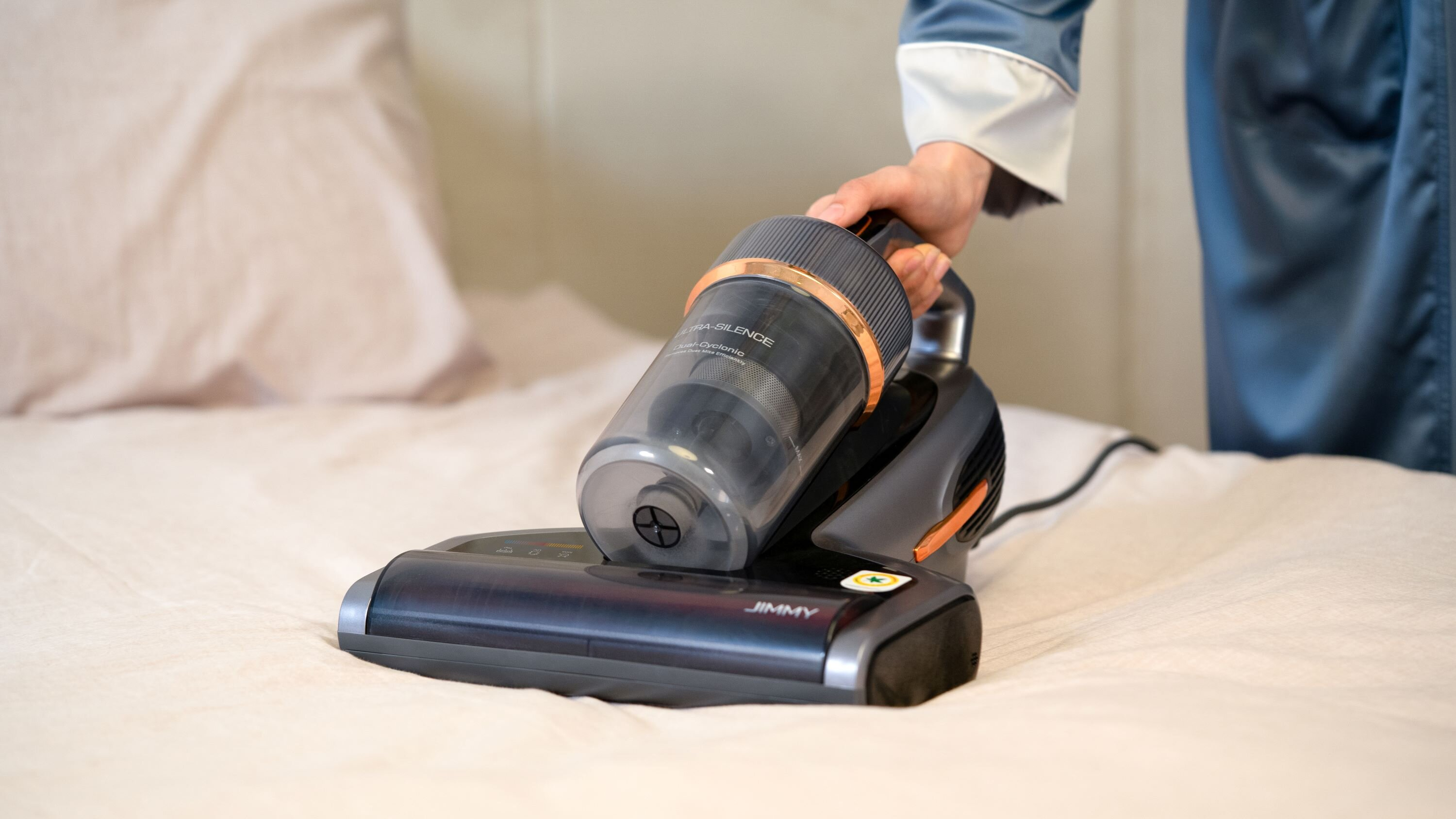 Osoba trzyma bezprzewodowy odkurzacz na łóżku. Odkurzacz jest czarny i miedziany, widoczna jest marka Jimmy.