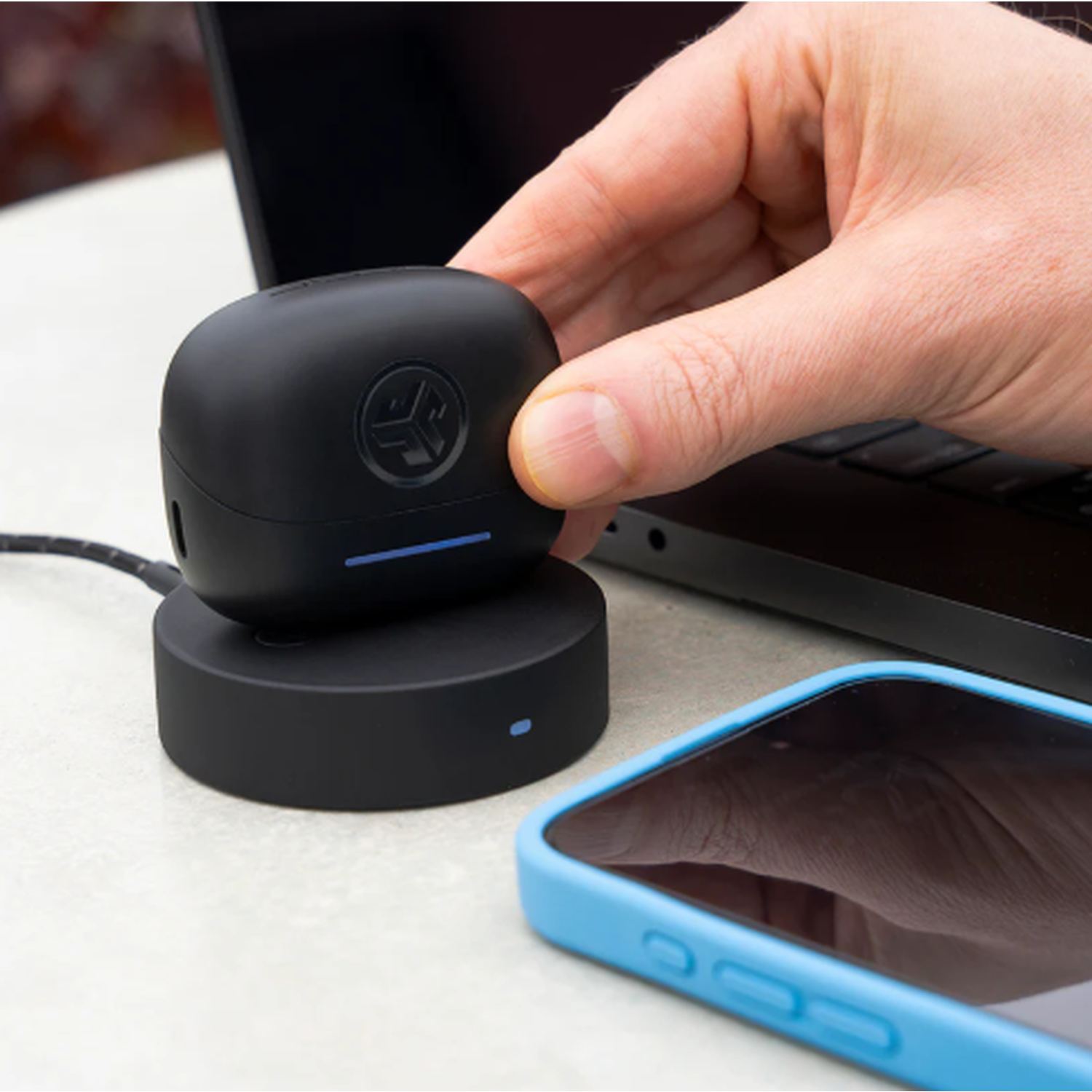 Een hand plaatst een zwarte oortelefoonhoes op de zwarte oplaadbasis. Een telefoon met blauwe hoes en een laptop staan ook op de tafel.