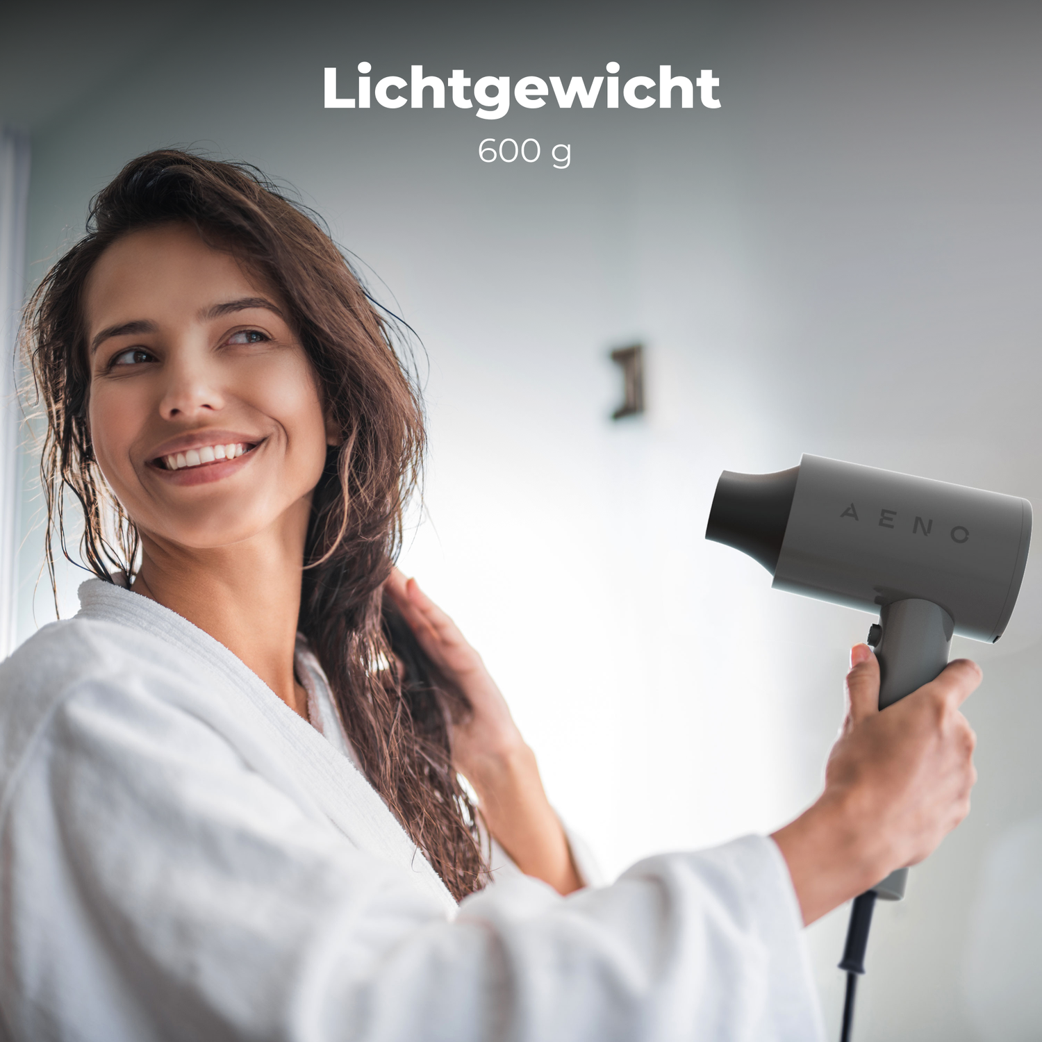 A woman in a white robe smiles, holding a hairdryer. The hairdryer is gray, with the brand AENO on it. The background is blurry.