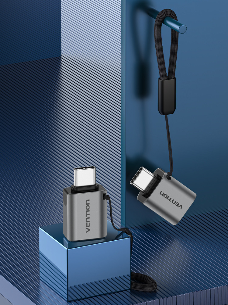 Két Vention USB-C adapter. Az egyik egy kék kockán nyugszik, a másik lóg. Mindkettő ezüst színű, fekete pántokkal és sötétkék háttérrel.
