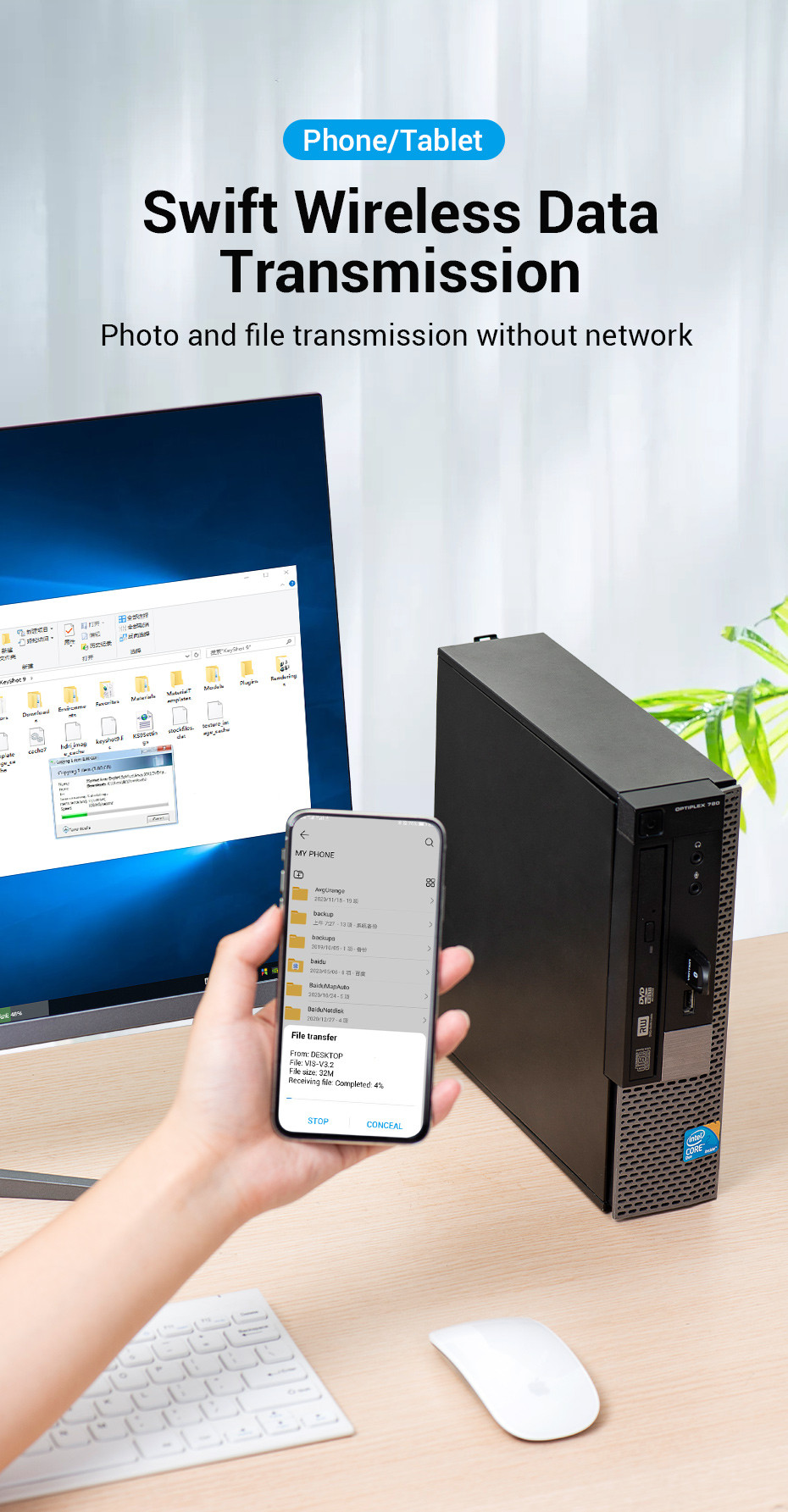 Egy kéz egy telefont tart, amely fájlátvitelt mutat. A háttérben egy asztali számítógép és monitor. Egér és billentyűzet az előtérben.