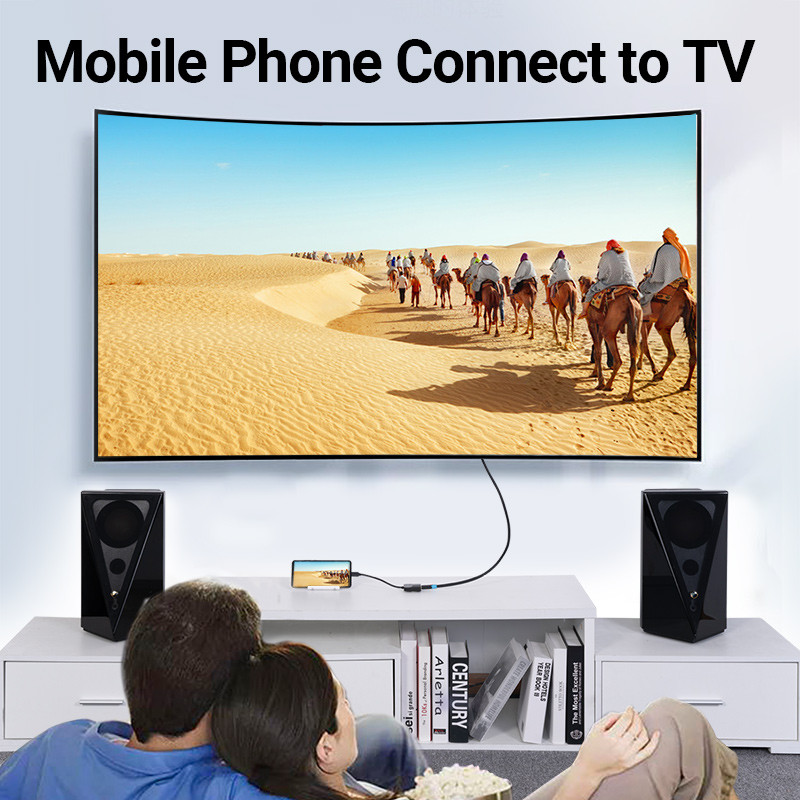 Egy pár egy TV-t néz, amely egy sivatagi jelenetet mutat tevékkel. A telefon a TV-hez csatlakozik. A hangszórók a TV-t szegélyezik. Könyvek és pattogatott kukorica láthatók.