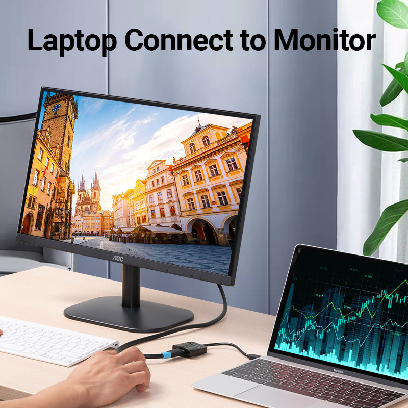 Egy laptop egy monitorhoz van csatlakoztatva. Egy kéz tart egy kábelt. A monitor egy városi jelenetet mutat. A laptop egy grafikon.