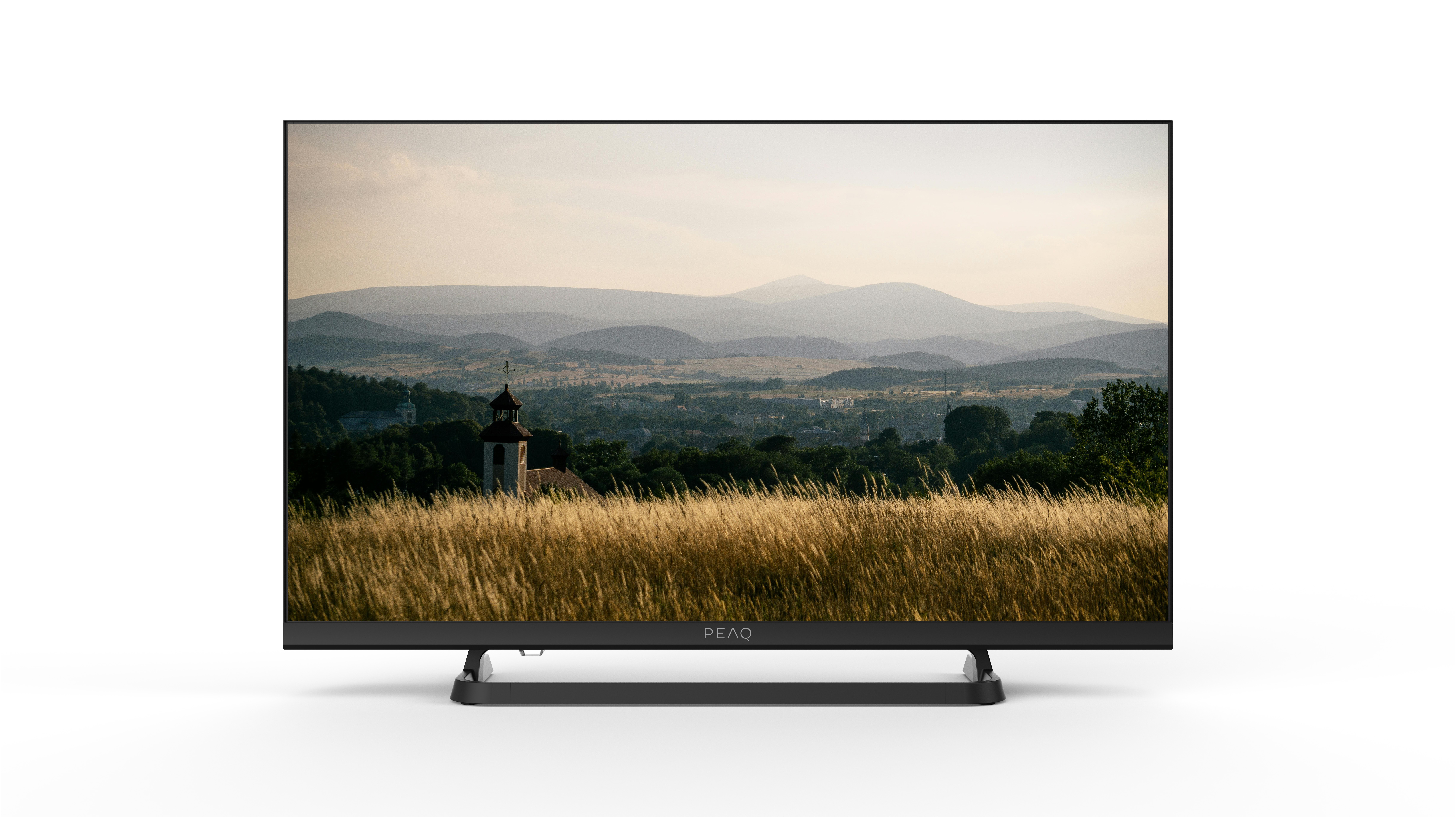 Telewizor wyświetla krajobraz z górami, miastem i wysoką trawą. Telewizor jest czarny i stoi na białej powierzchni.