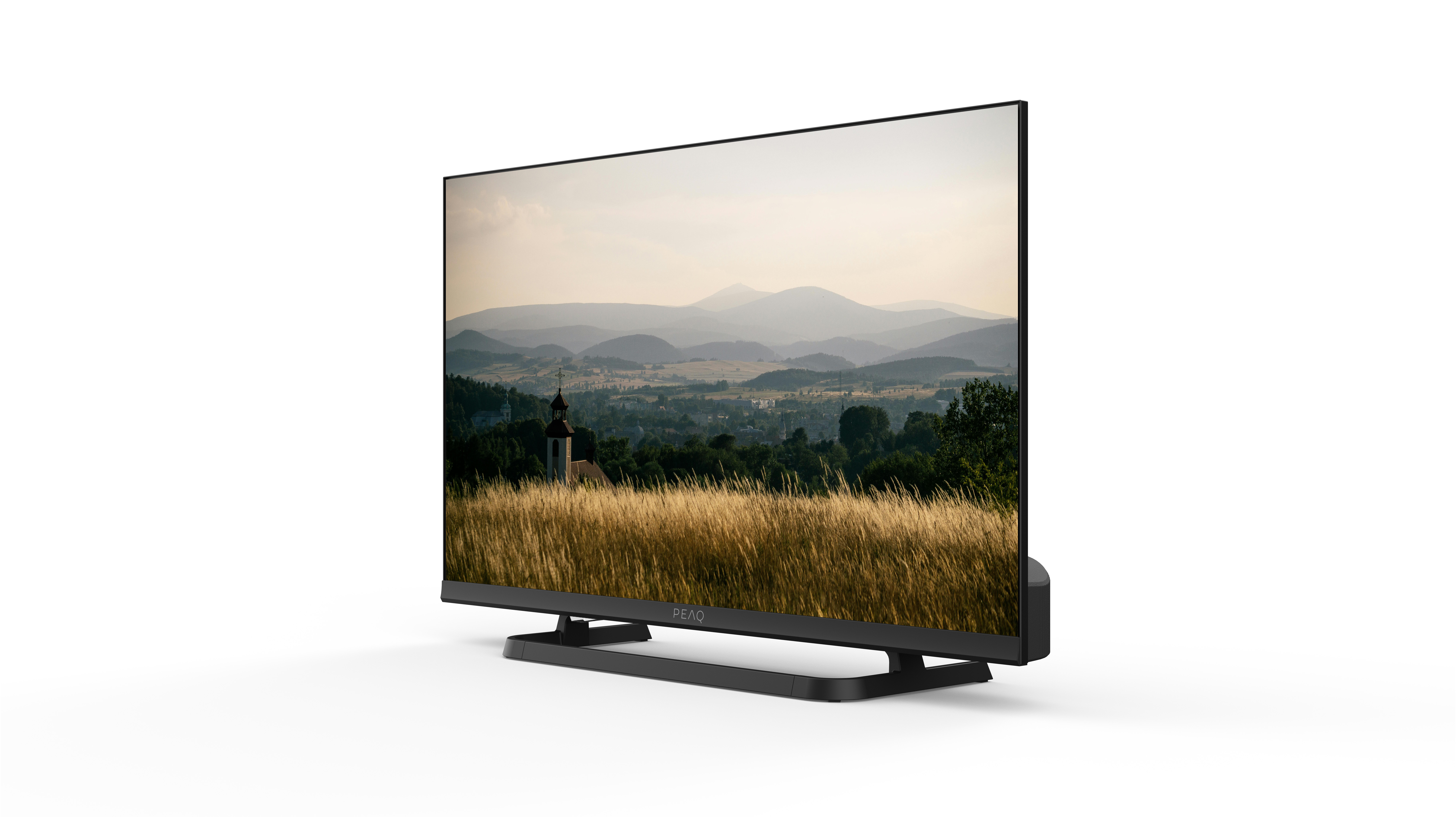 Telewizor wyświetla krajobraz z górami, kościołem i wysoką trawą. Telewizor jest czarny i stoi na białej powierzchni.