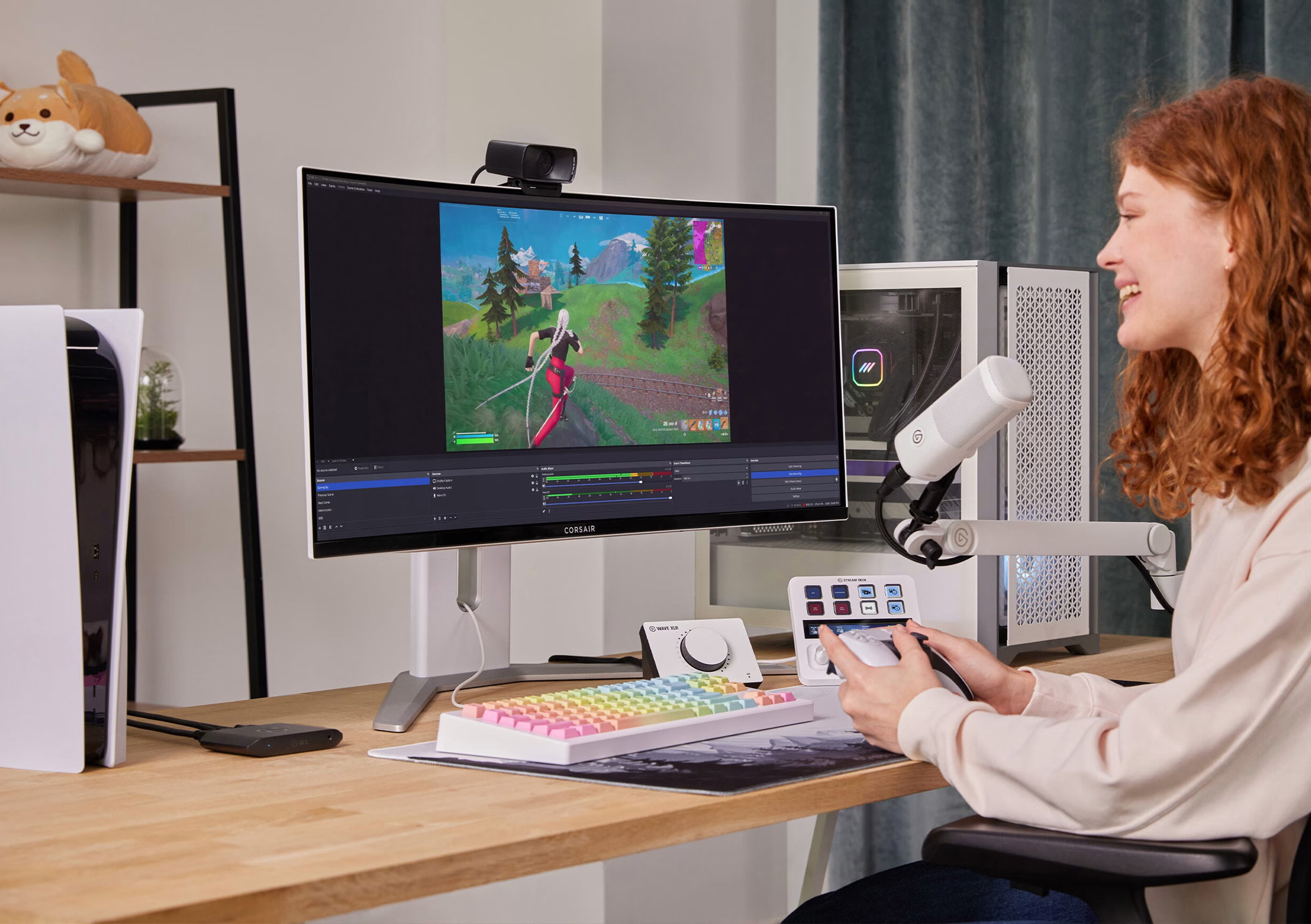 Eine Frau streamt ein Videospiel auf einem Monitor. Tastatur, Maus und Mikrofon sind sichtbar. Eine PS5-Konsole im Hintergrund.