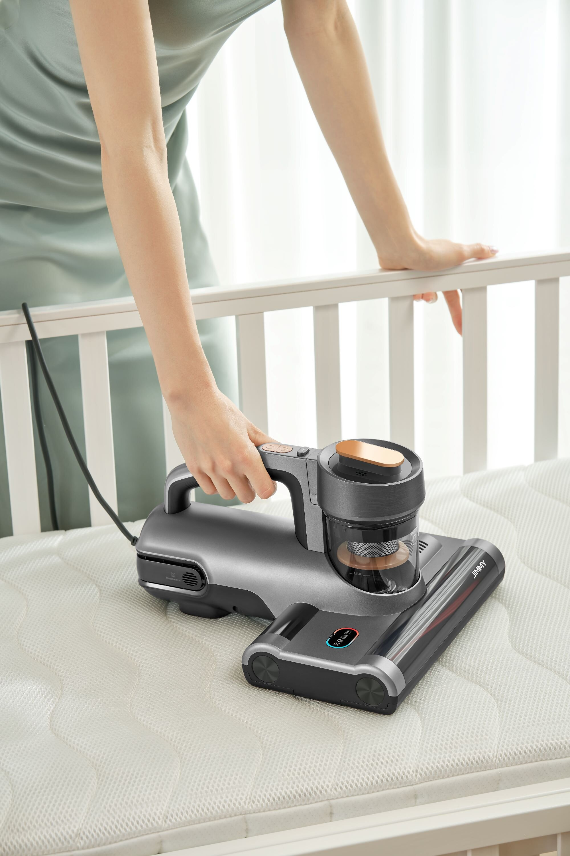 Osoba używa szarego odkurzacza ręcznego na białym materacu. Odkurzacz ma złoty akcent. Tło to białe łóżeczko.