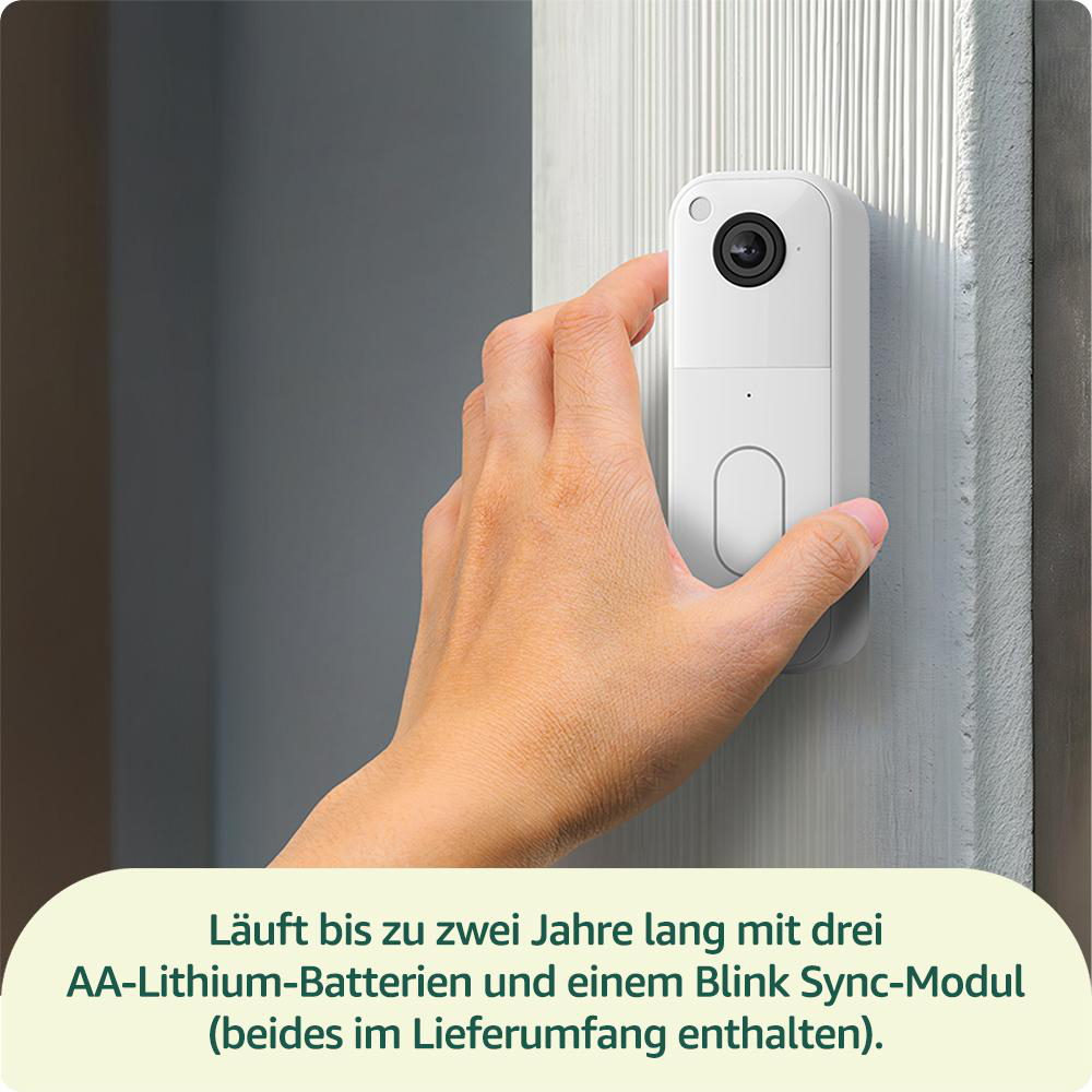 Eine Hand hält eine weiße Türklingel mit Kamera. Die Türklingel ist an einer hellen Wand montiert.