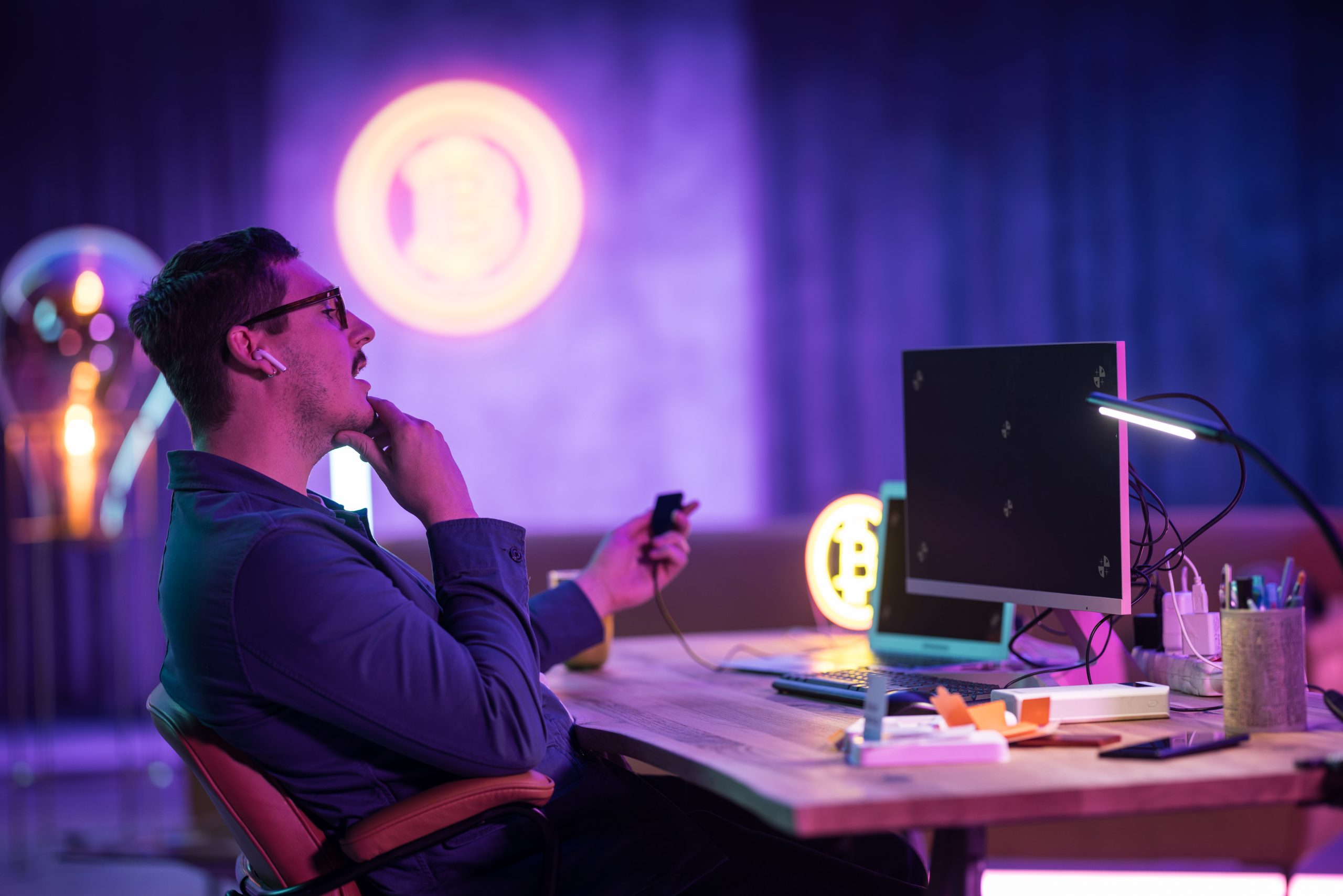 Mężczyzna w okularach i Airpods przy biurku. Znaki kryptowalut w tle. Monitory, lampa i telefon na biurku.