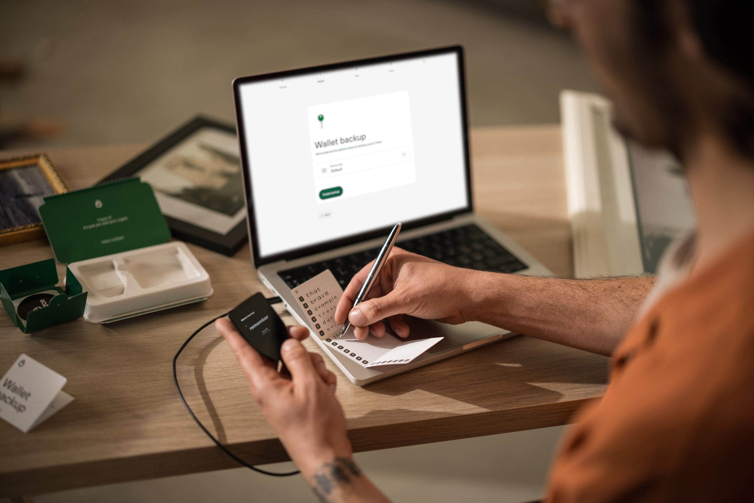 Osoba korzysta z laptopa i pisze na notatniku. Urządzenie jest podłączone do laptopa. Inne przedmioty leżą na drewnianym stole.