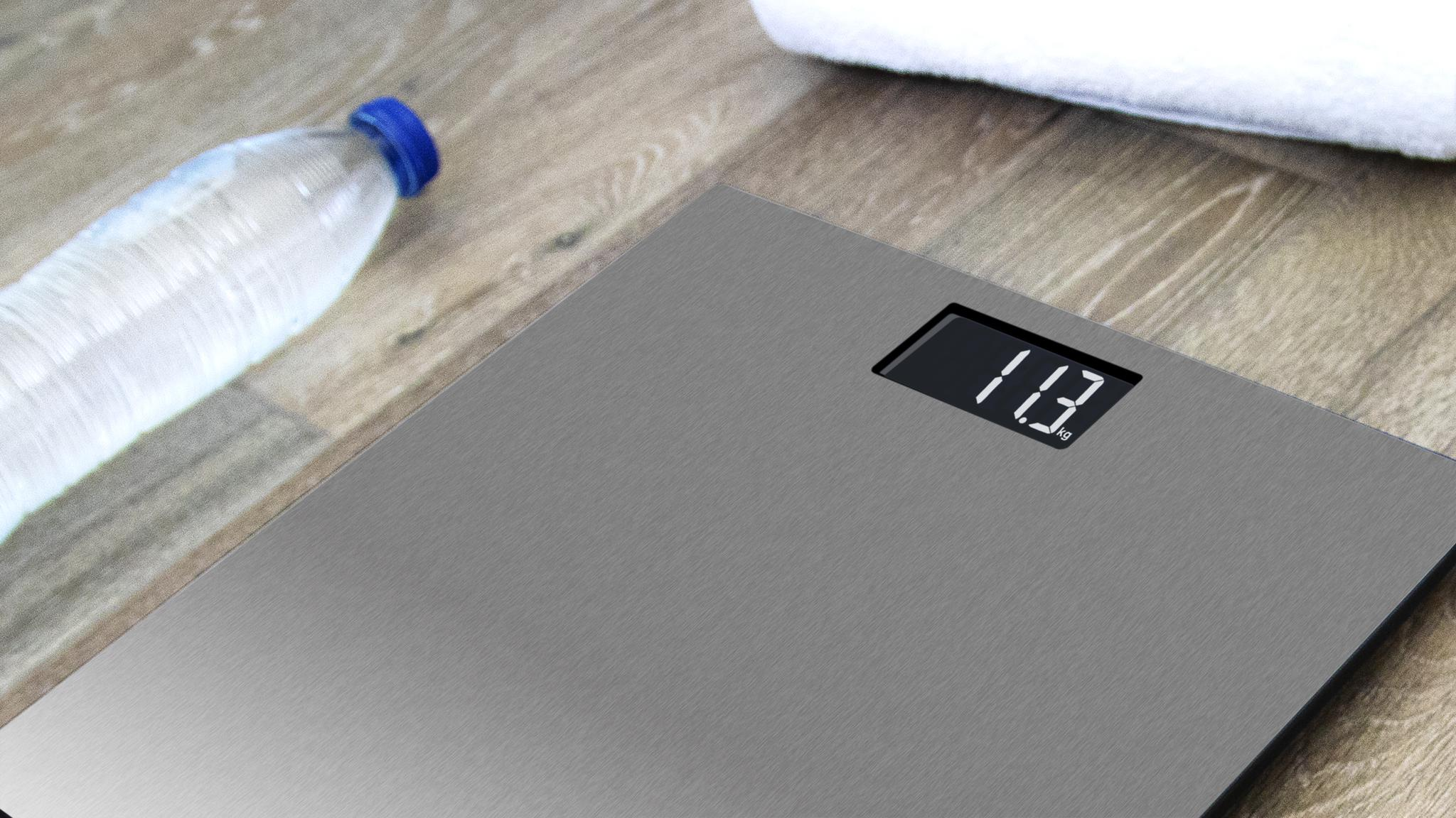 Una báscula digital muestra 11.3 kg. Una botella de agua con tapa azul y una toalla blanca en madera.
