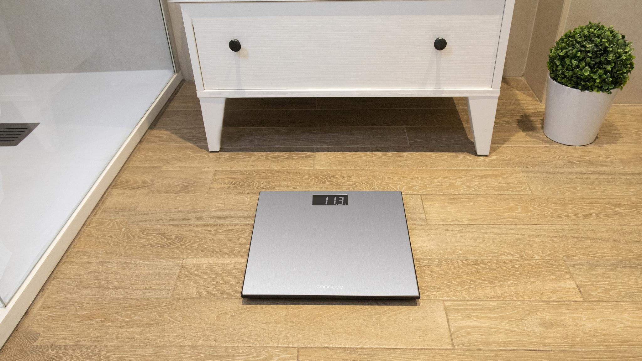 Una escena de baño con una báscula digital, un mueble blanco y una planta en una maceta blanca. El suelo es de madera.