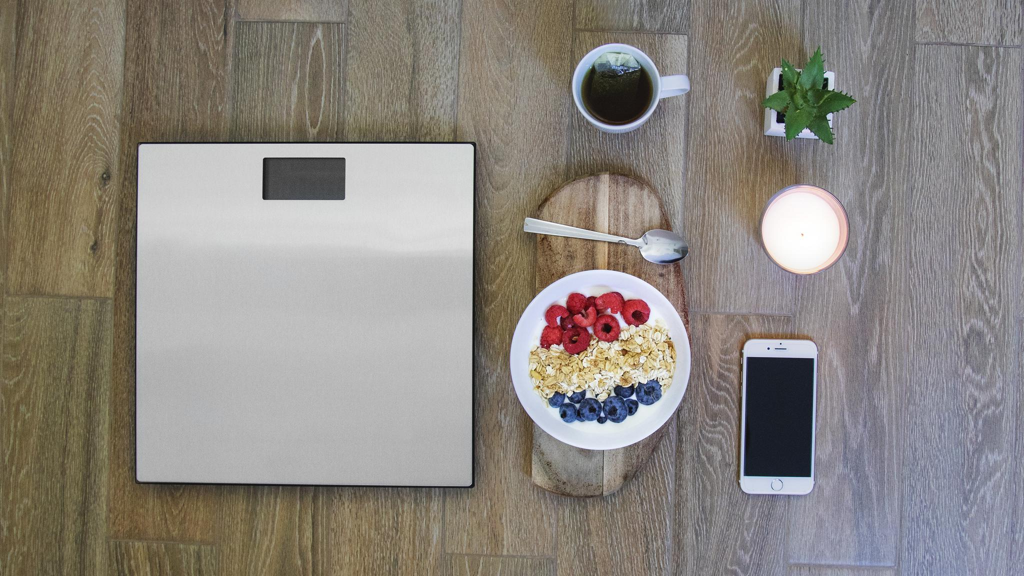 Vista superior: balanza, cuenco de desayuno con bayas, taza de té, teléfono, planta, vela sobre suelo de madera.