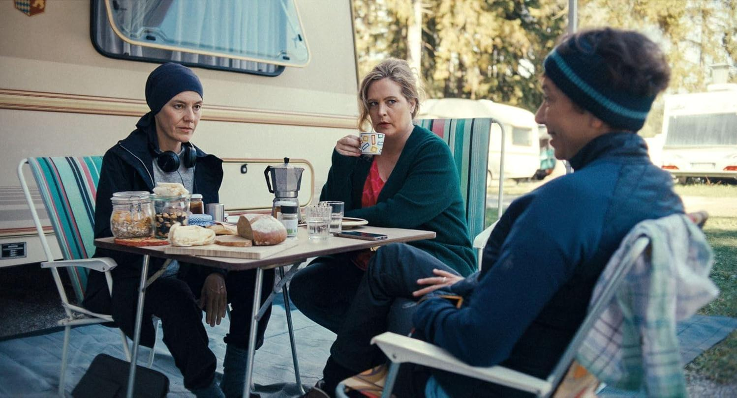 Drei Personen sitzen an einem Tisch vor einem Wohnwagen. Essen und Kaffee stehen auf dem Tisch. Eine Frau trägt ein Kopftuch, eine andere trinkt Kaffee.