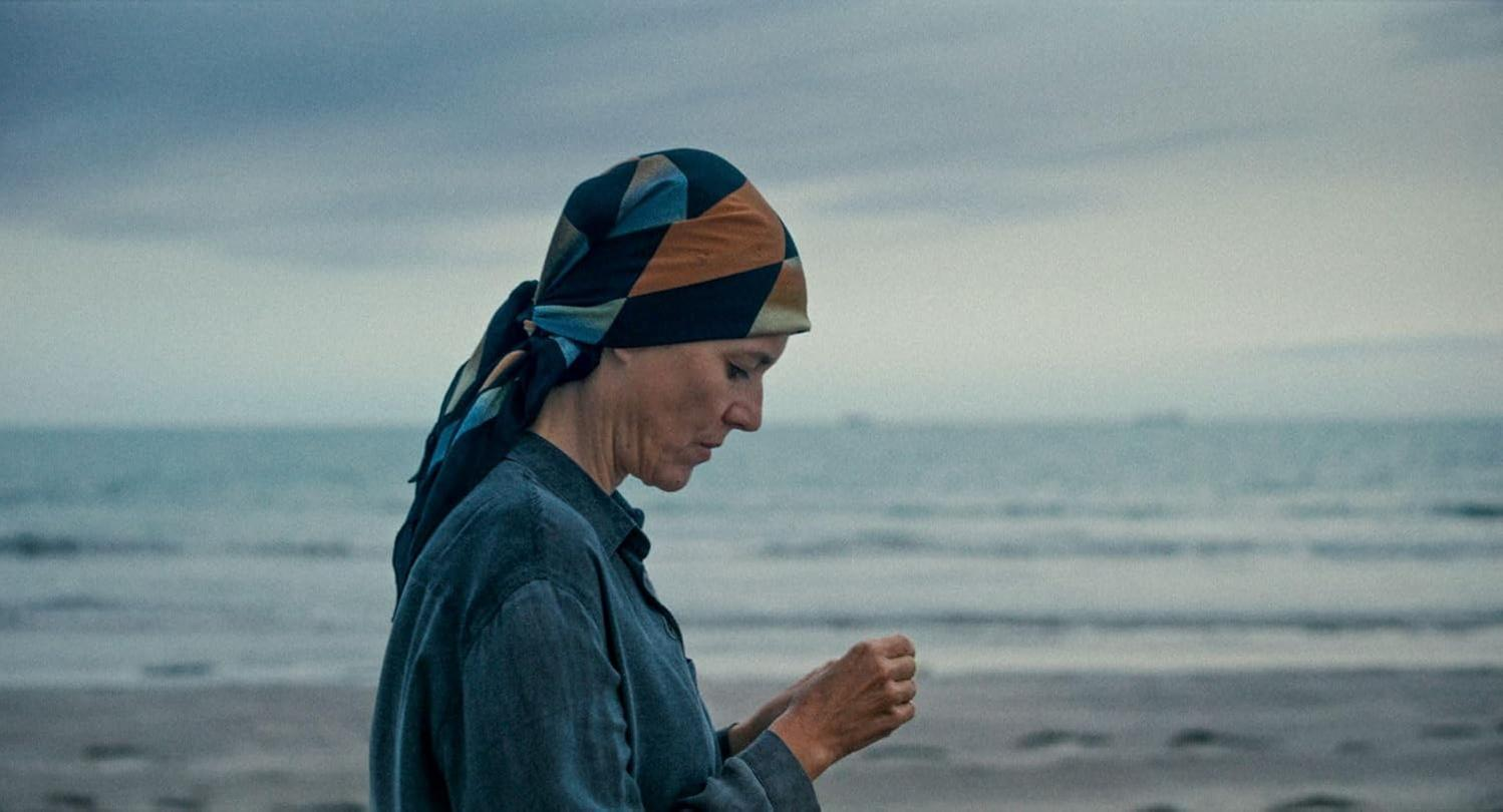 Eine Frau mit Kopftuch steht am Strand und blickt nach unten. Im Hintergrund sind Meer und Himmel, was eine gedrückte Stimmung erzeugt.