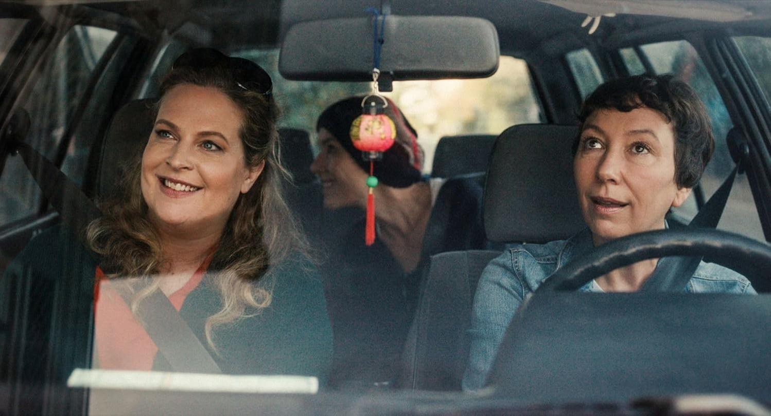 Drei Frauen in einem Auto. Eine lächelt, eine fährt und eine sitzt hinten. Eine rote Laterne hängt am Rückspiegel.
