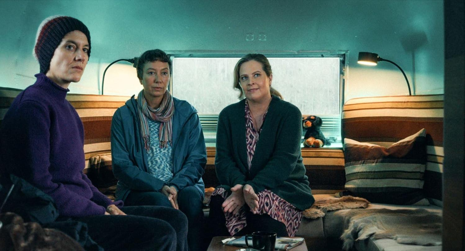 Drei Frauen sitzen in einem Wohnwagen. Eine trägt einen lila Pullover und eine Mütze. Eine andere trägt eine blaue Jacke und die dritte eine grüne Strickjacke.
