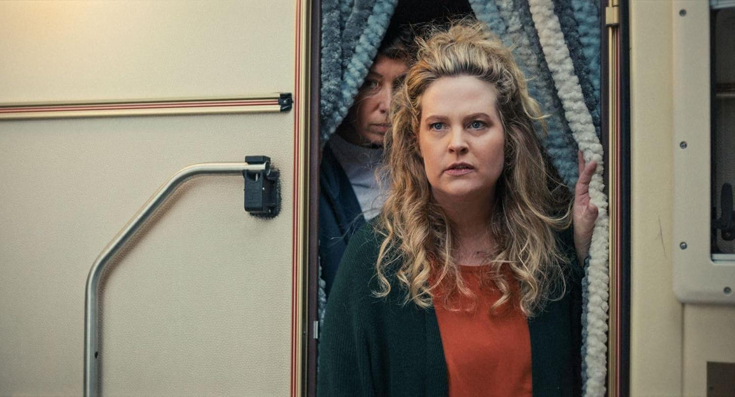 Zwei Frauen in einer Wohnmobiltür. Eine mit blonden Haaren schaut nach vorne, die andere blickt hinter einem Vorhang hervor. Beide haben ernste Ausdrücke.