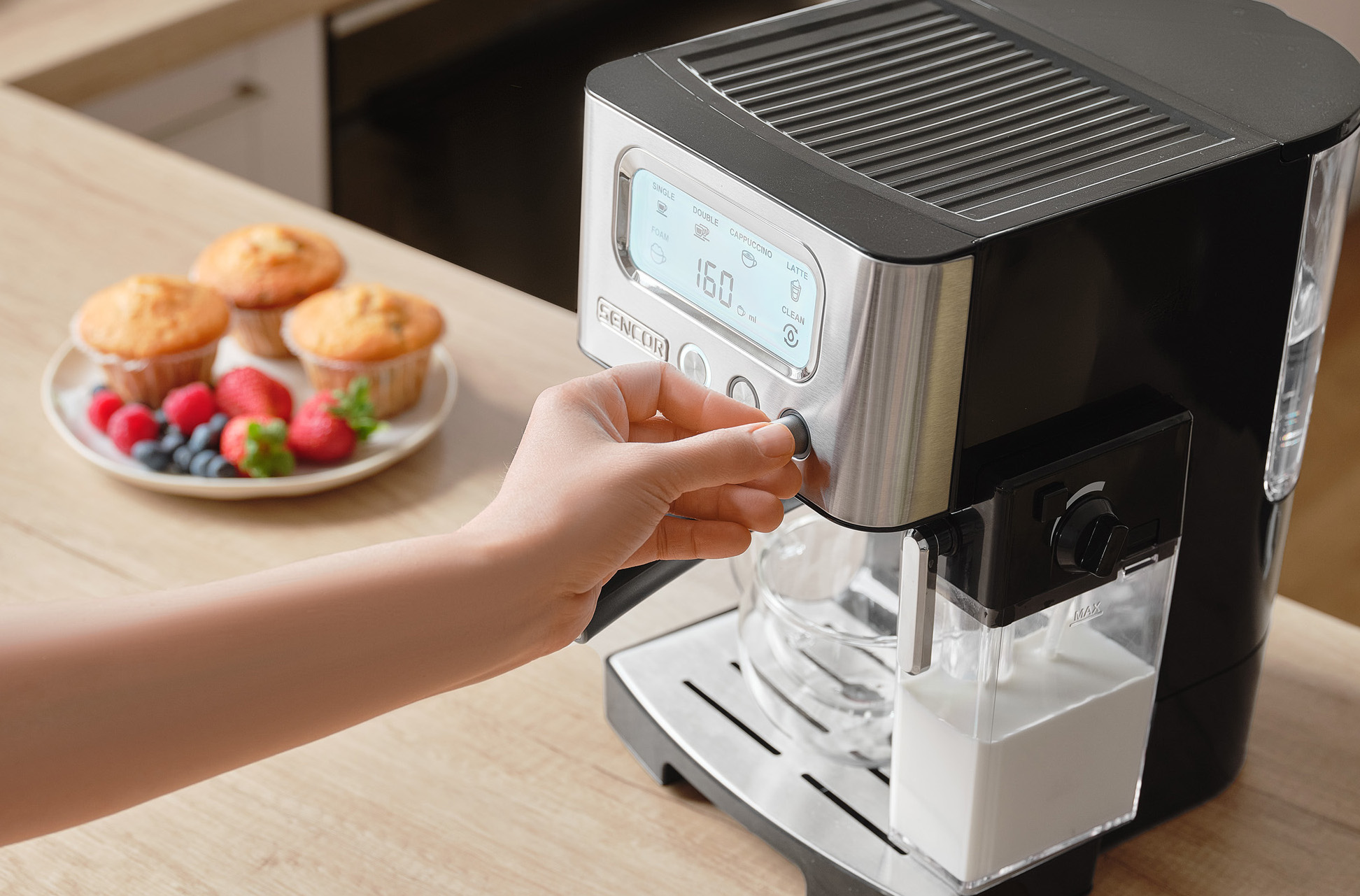 Ręka obsługuje ekspres do kawy. Muffiny i jagody leżą na talerzu. Maszyna ma cyfrowy wyświetlacz i pojemnik na mleko.