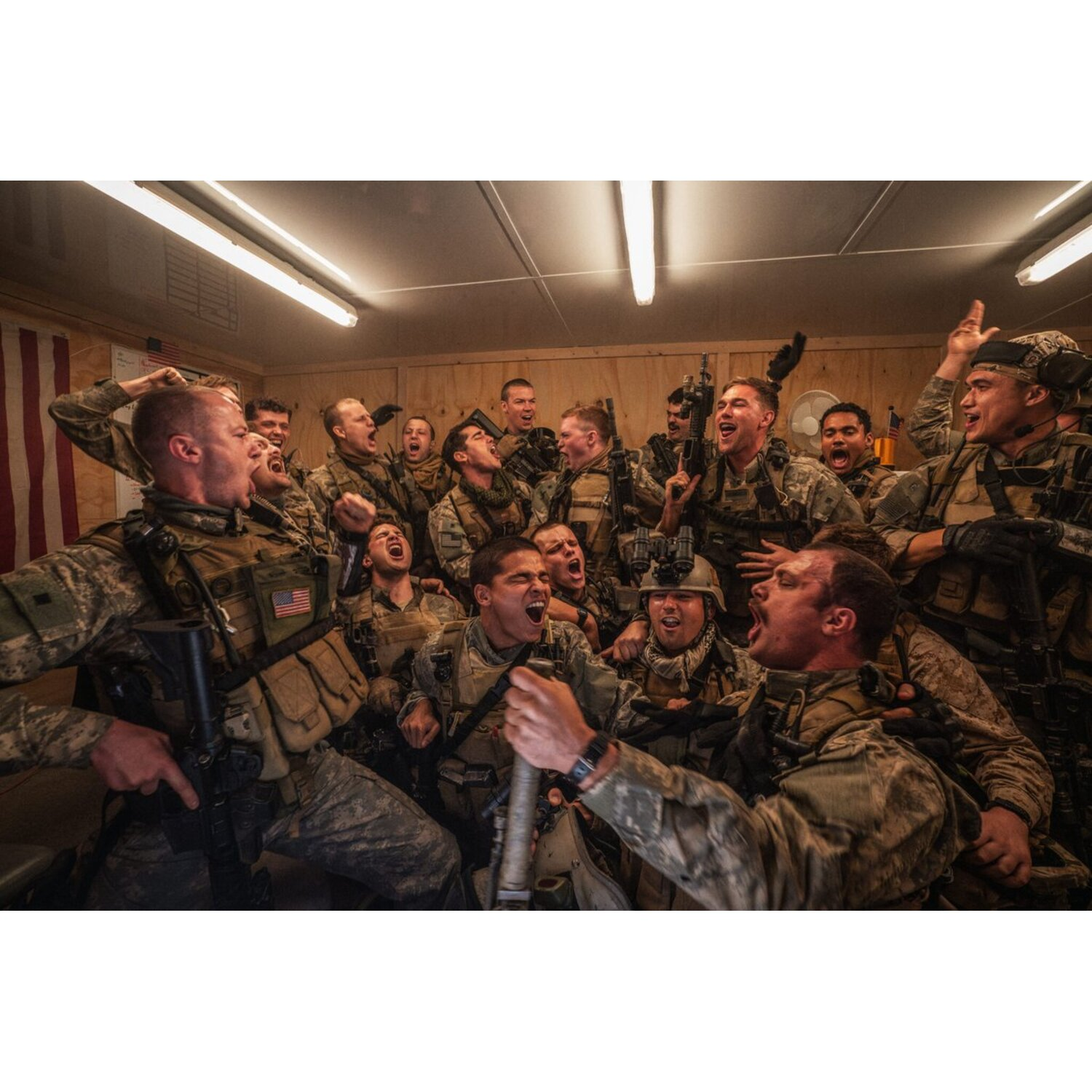 Group of soldiers celebrating indoors, many shouting. They wear camouflage uniforms and tactical gear. An American flag hangs in the background.
