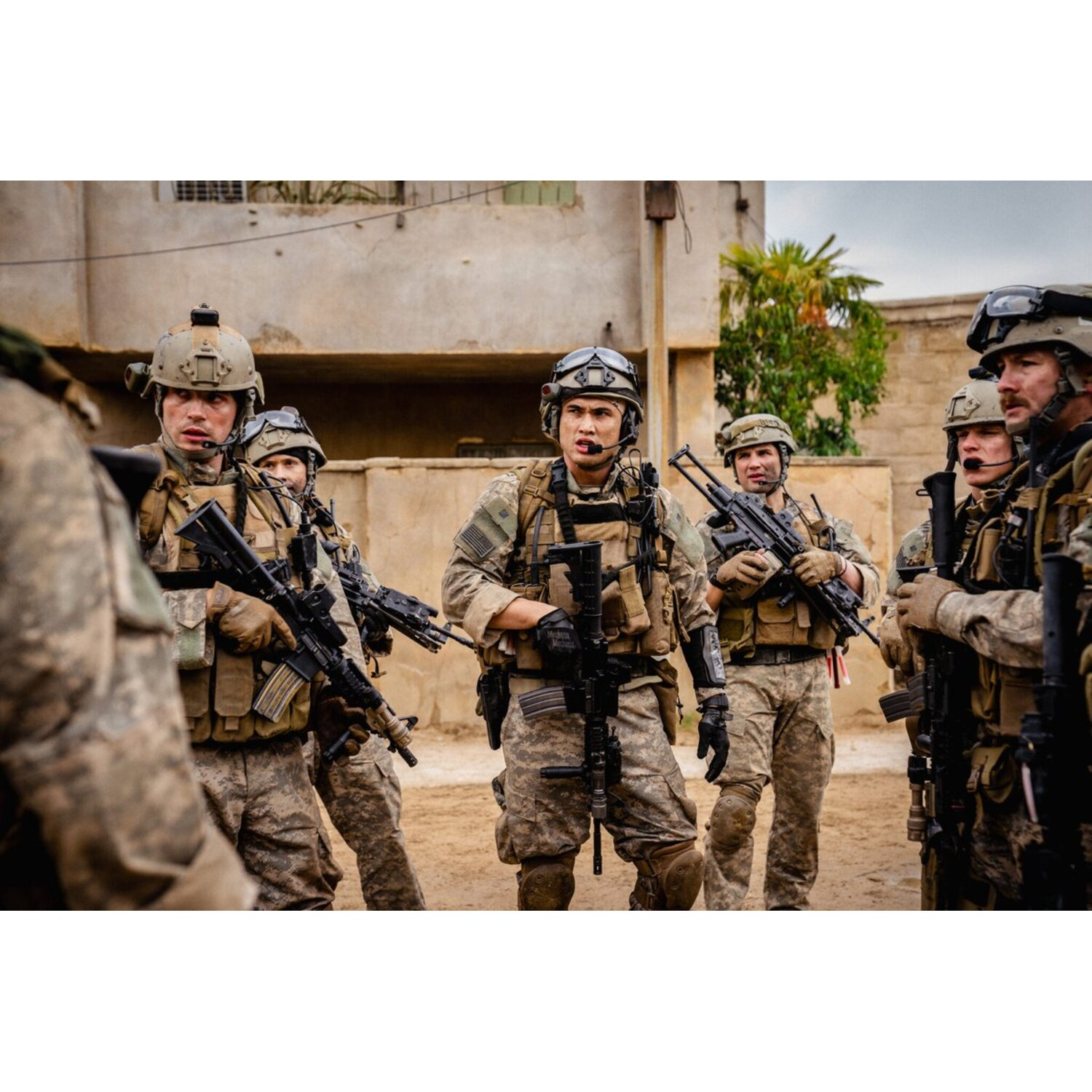 Group of soldiers in combat gear, holding rifles, standing in a sandy area near buildings and a palm tree. Cloudy sky in the background.