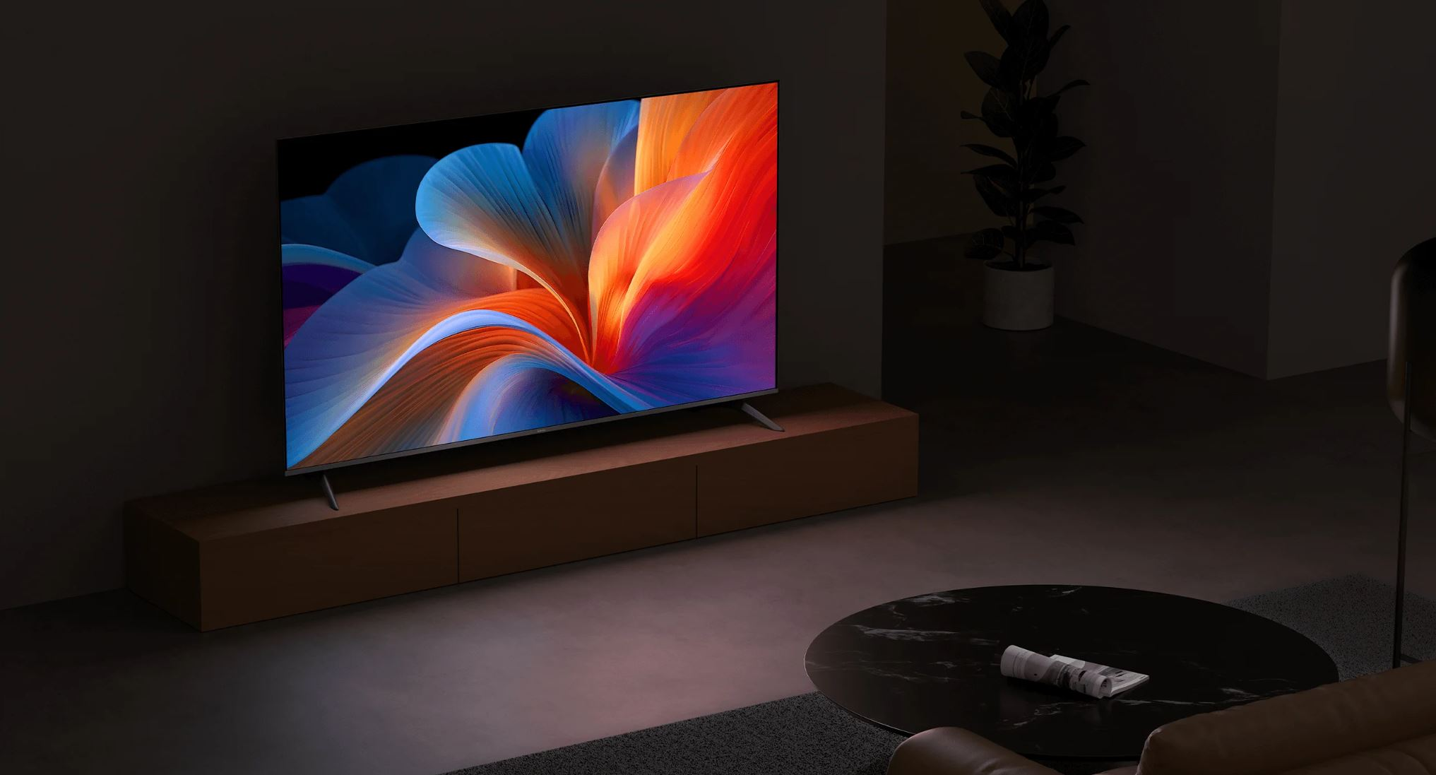 Duży telewizor wyświetla kolorową sztukę abstrakcyjną. Stoi na drewnianej szafce w słabo oświetlonym pomieszczeniu. Widoczna jest roślina i stolik kawowy.