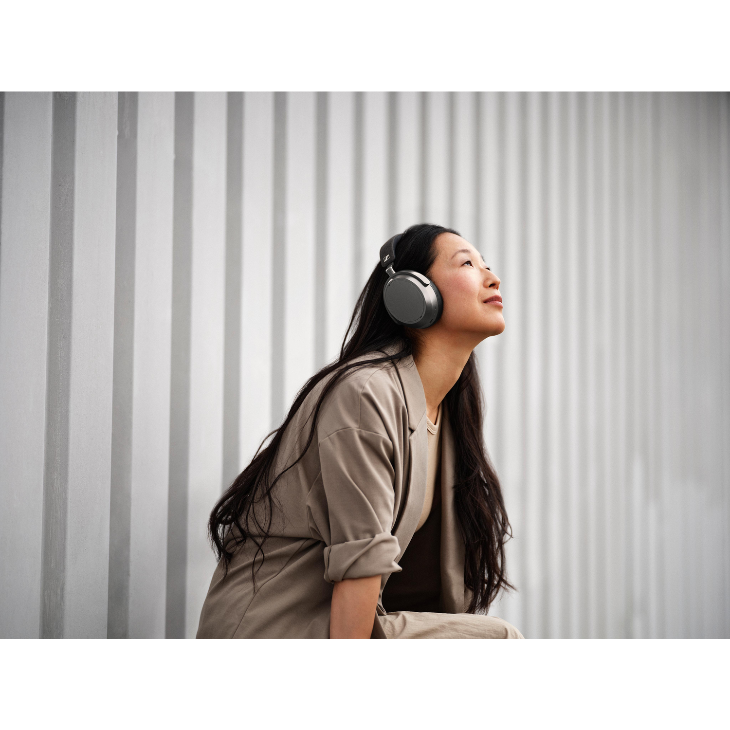 A woman with long black hair wearing headphones looks up. She's in a beige suit, sitting in front of a textured white wall.