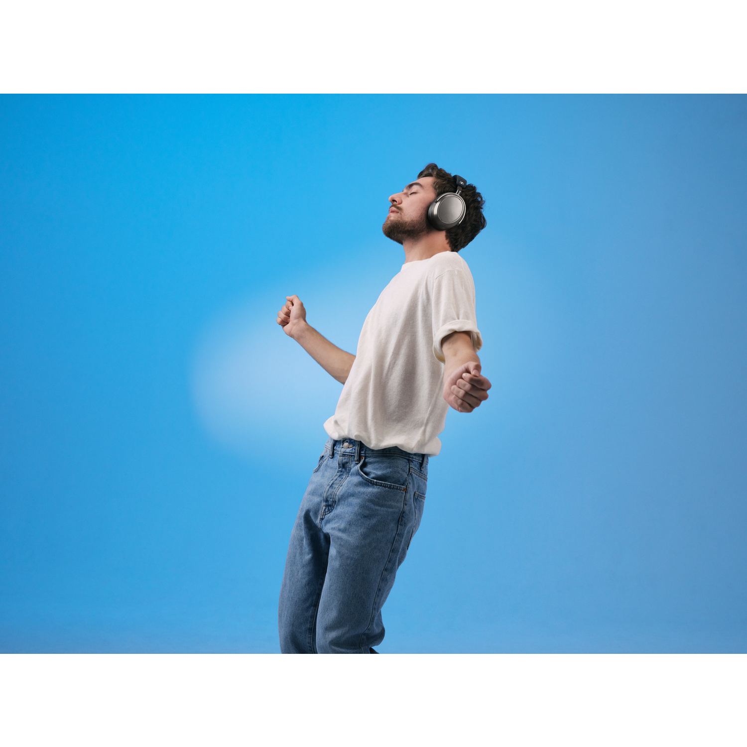 A man wearing headphones, a white t-shirt, and blue jeans stands with arms outstretched, eyes closed, against a blue background.
