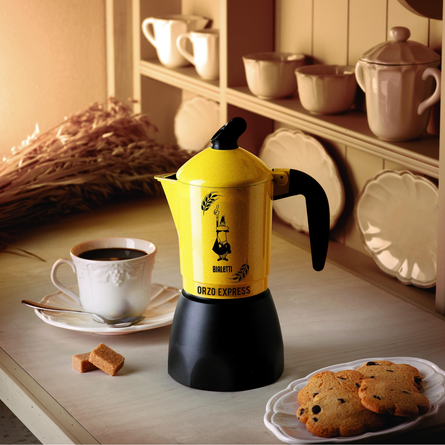 Una caffettiera gialla con base nera, una tazza di caffè, biscotti e zollette di zucchero su un tavolo. Sfondo: mensole con tazze e piatti.