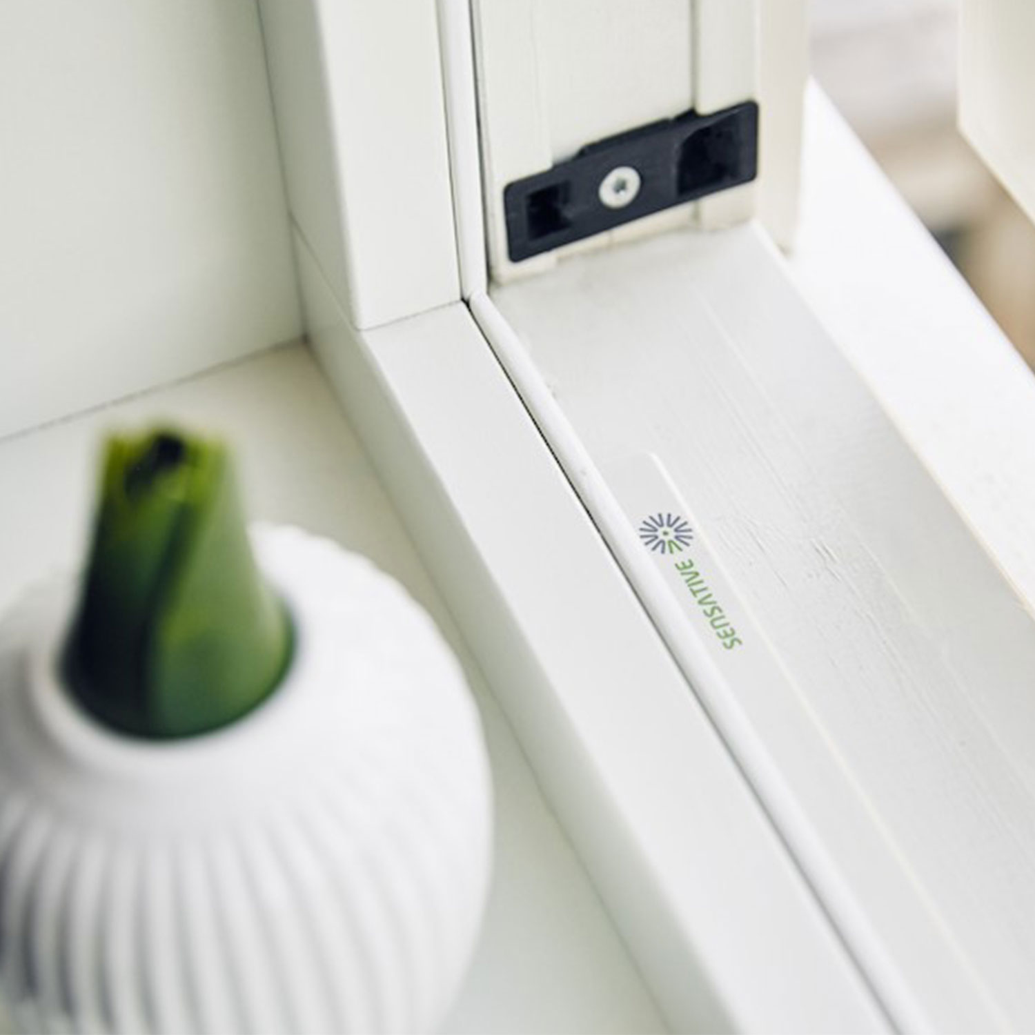Weißer Fensterrahmen mit schwarzem Riegel, weiße Vase mit grünem Aufsatz und ein mit 'SENSITIVE' beschrifteter Stab.