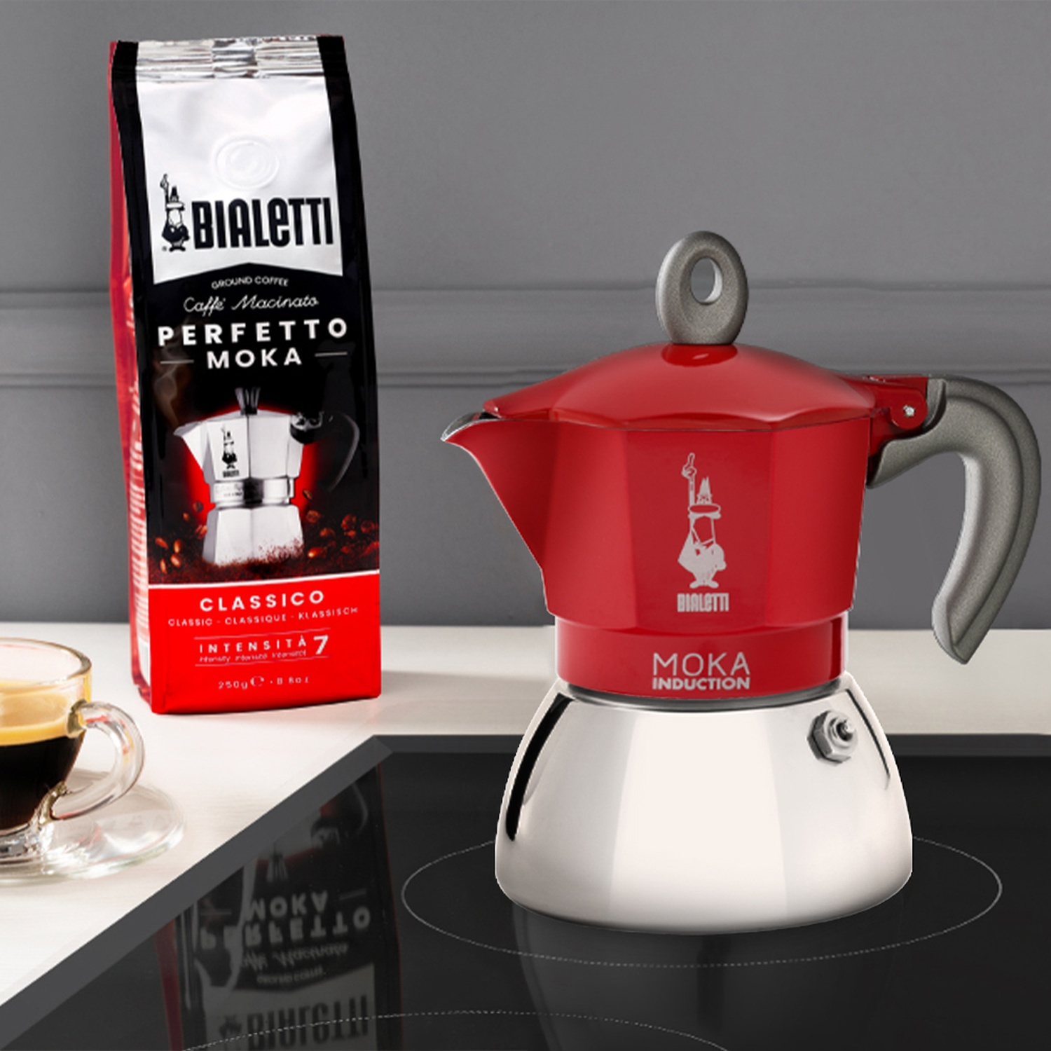 Caffettiera Moka Bialetti rossa su un piano cottura nero con una confezione di caffè e una tazza di caffè.