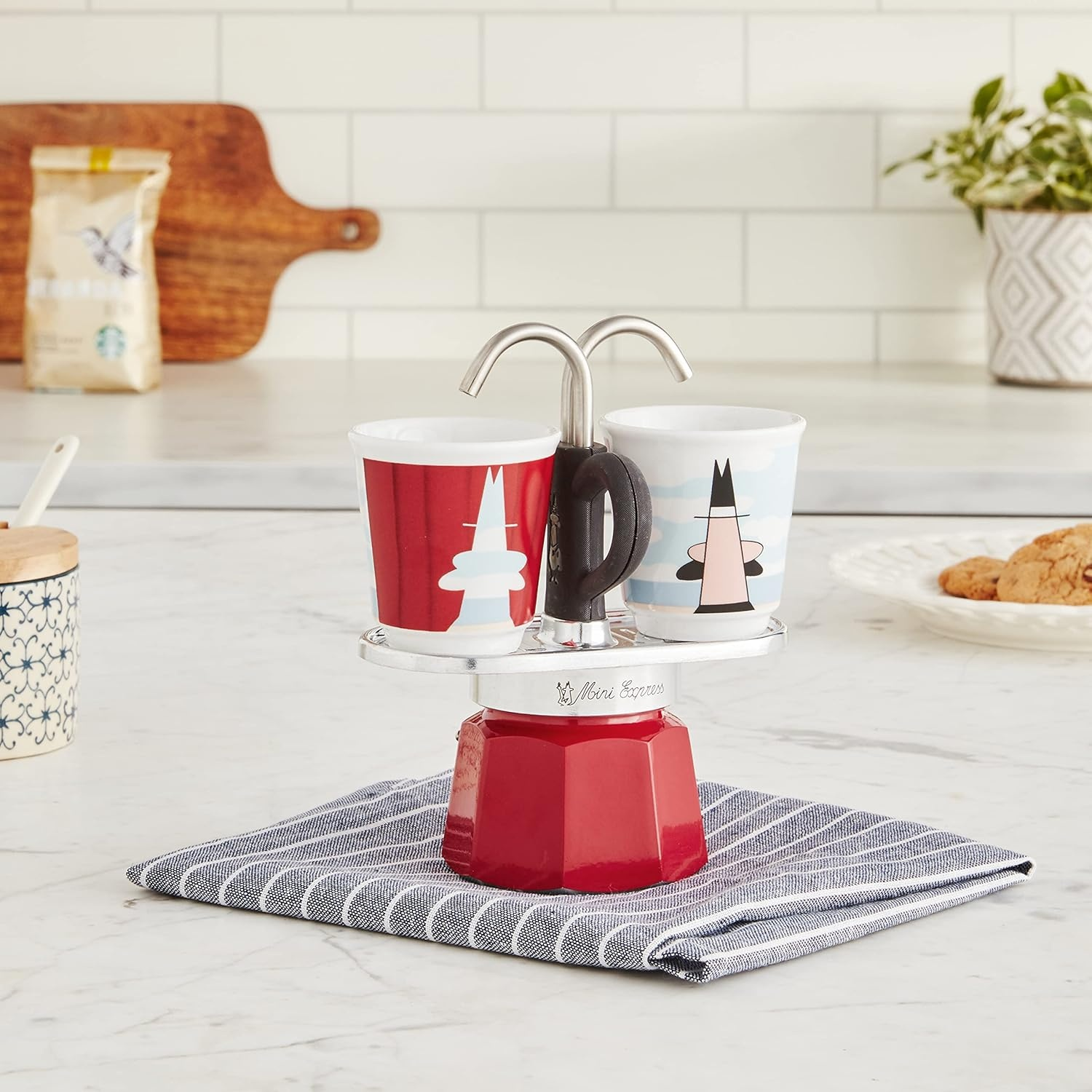 Una caffettiera rossa con due tazze su un panno a righe. Un sacchetto di caffè e biscotti sullo sfondo.